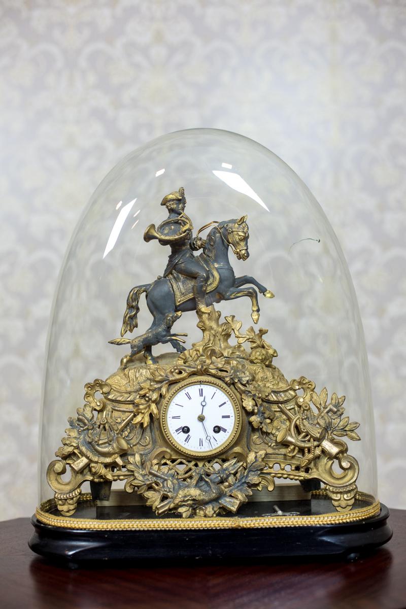 A clock with a glass shade, circa 1930, on a wooden base.
The striking, metal casing of the clock is decorated with hunting emblems, and is crowned with a semi-plastic figure of a huntsman on a horse.
Presented clock can be winded with a key. It