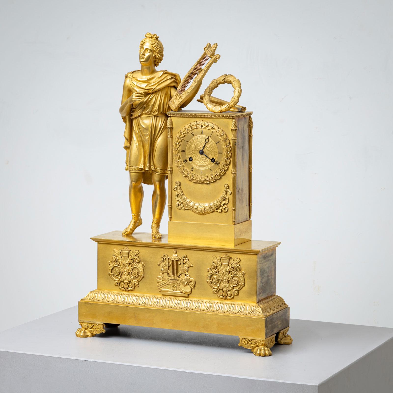 Large fire-gilded bronze pendulum clock on lion's paws with a rectangular pedestal featuring lyre and torch motifs. Above it rises the full-figure depiction of the god Apollo with a lyre and victory wreath. The clock mechanism is housed in a