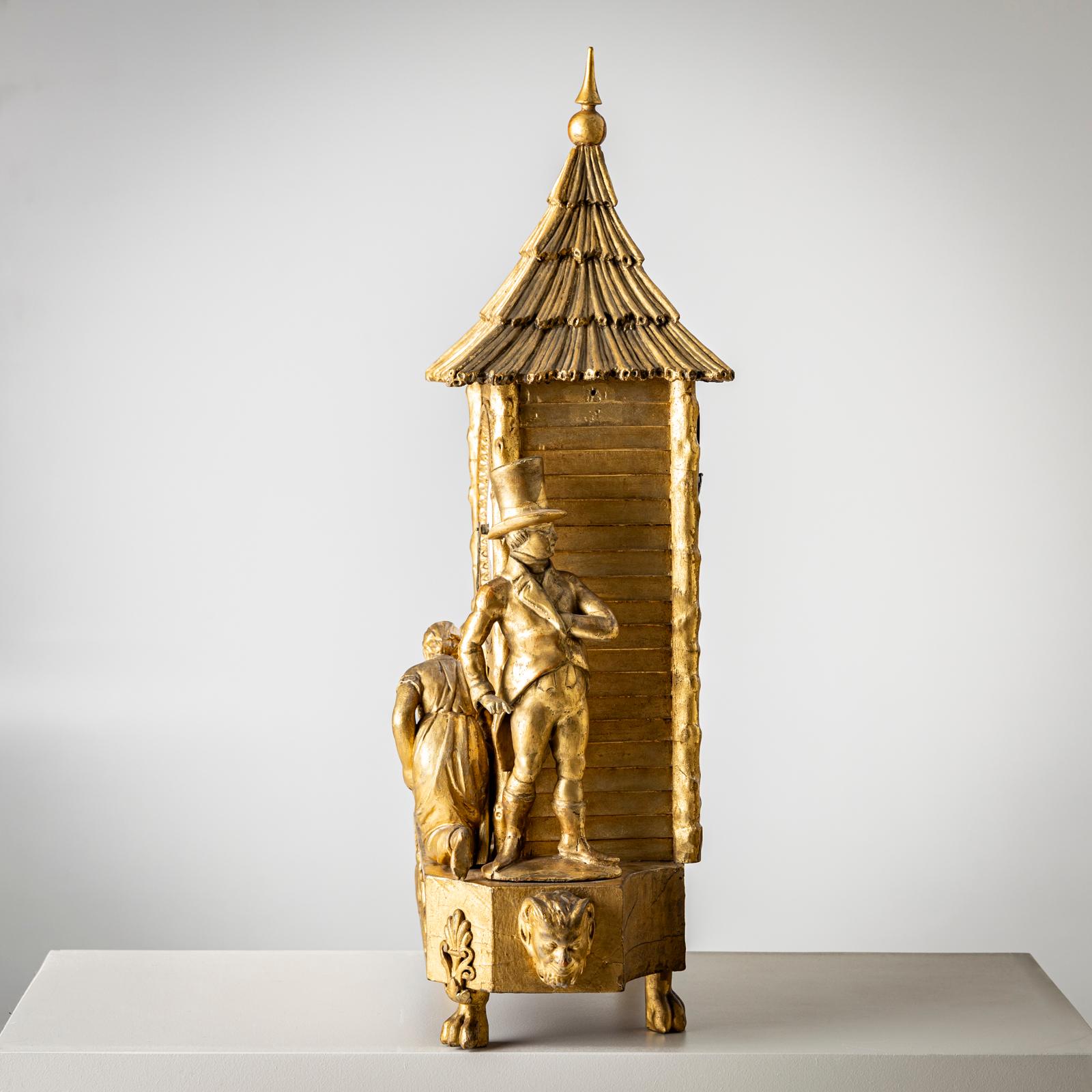 Kaminsimsuhr in einem goldpatinierten Holzgehäuse mit emailliertem Zifferblatt und achteckigem Sockel mit Tatzenfüßen. Darüber erhebt sich das Uhrengehäuse in Form einer Holzhütte mit einem spitzen Strohdach. Eine junge Frau füttert Vögel, links ist