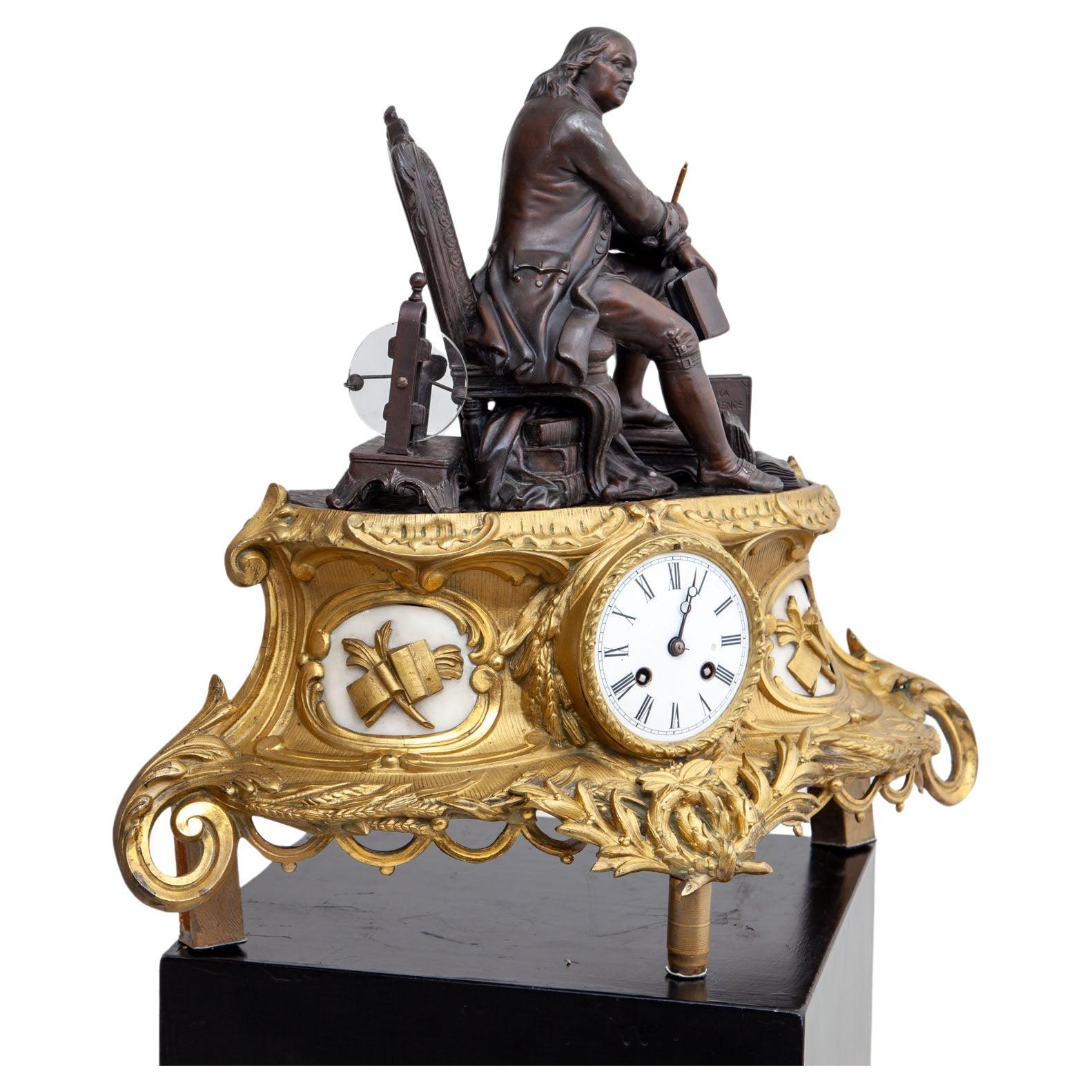 Große Kaminuhr aus Bronze mit vergoldetem Sockel mit Rocaille-Dekor, Kartuschen mit Schriftrollen und Büchern sowie einem Emailzifferblatt mit römischen Ziffern. Darauf ist aus brünierter Bronze ein sitzender Benjamin Franklin umgeben von Büchern