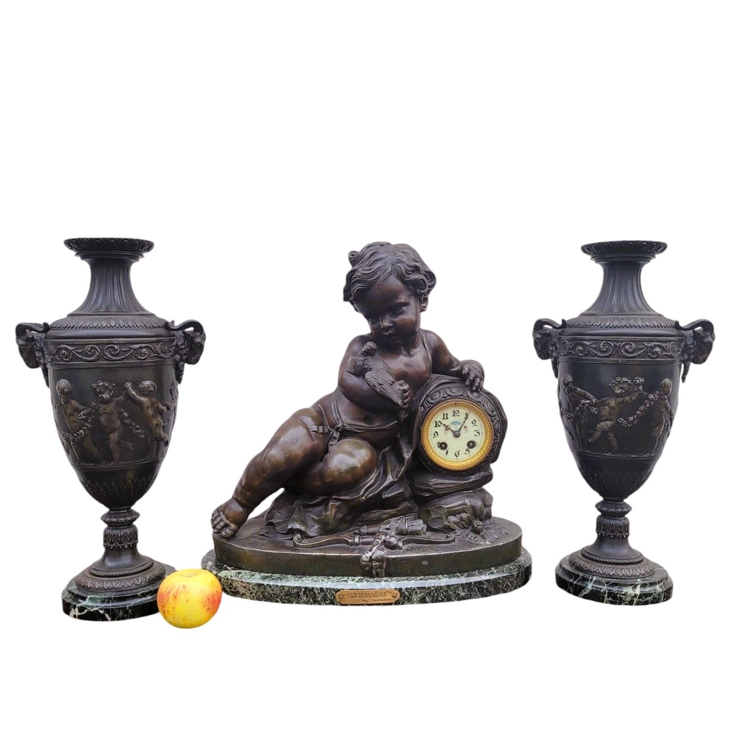 Kaminbesteck bestehend aus 3 Teilen, einer Uhr und 2 Vasen, alle aus patiniertem Zinn und grünem Marmor
Die Uhr ist mit einem Amor verziert, der zärtlich einen Vogel in den Armen hält und sich mit dem Zifferblatt an einen Felsen lehnt. Der Sockel