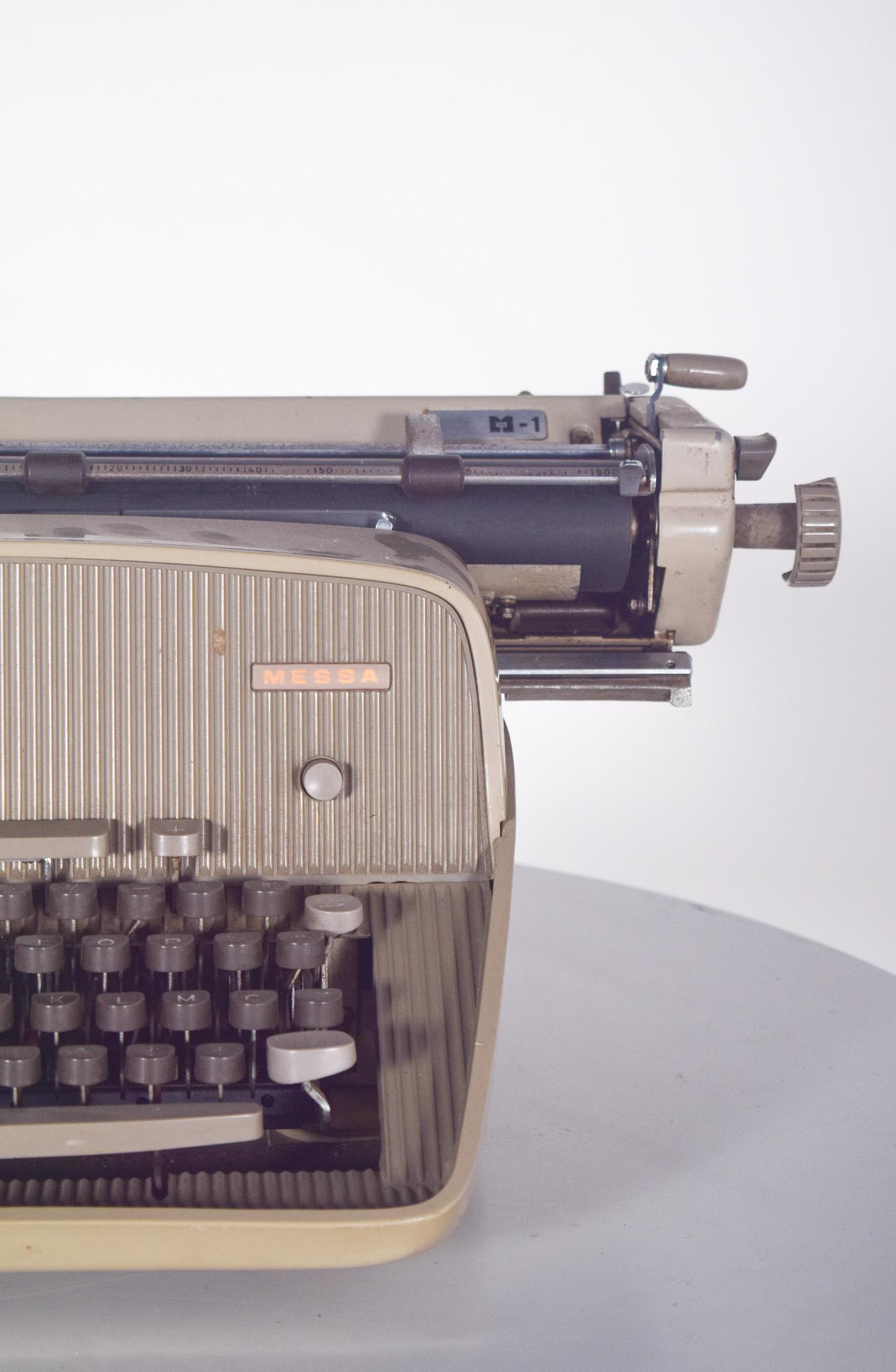 Machine à écrire manuelle, clavier AZERTY par MESSA. Avec un design robuste, une structure beige et des détails en métal chromé.
Le clavier comporte des touches rondes surélevées de couleur sombre, typiques des machines à écrire classiques, et le