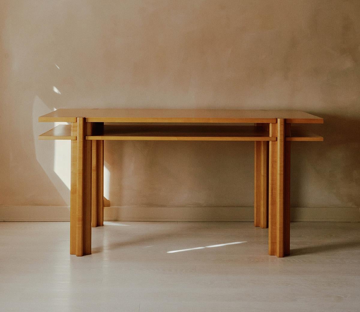 The Parallel desk is built around two suspended horizontal slabs that create an expansive workspace. The structure is anchored by cross-shaped legs that pierce through the top and frame a gap for open storage. The contrast between rounded edges and
