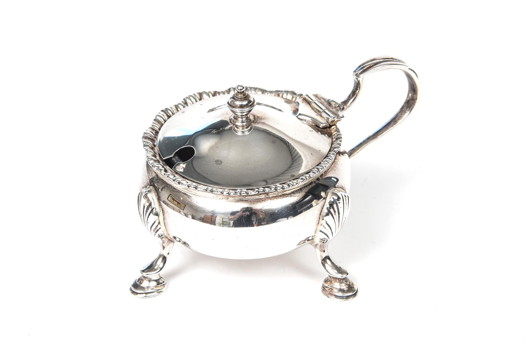 This beautiful ornate example of a mustard pot by famed Birmingham England silversmith Mappin and Webb was manufactured in 1929. The rim has a scolloped ribbon edge. It then bows out and sits on three ornate applied legs. It hinges open and has a
