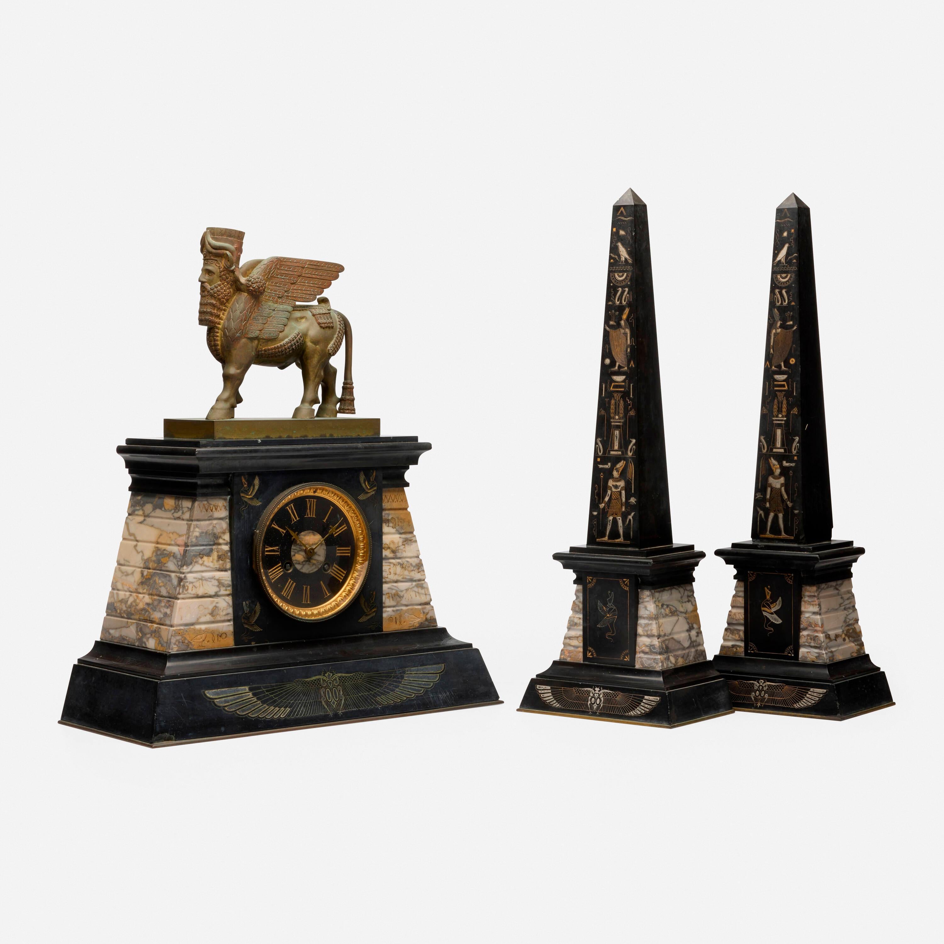 Marble and Patinated Bronze Egyptian Revival Mantel Garniture Set Set is comprised of a clock and a pair of obelisks. Clock features a bronze lamassu and engraved and gilt hieroglyphs. Impressed manufacturer's mark and number to clock movement.