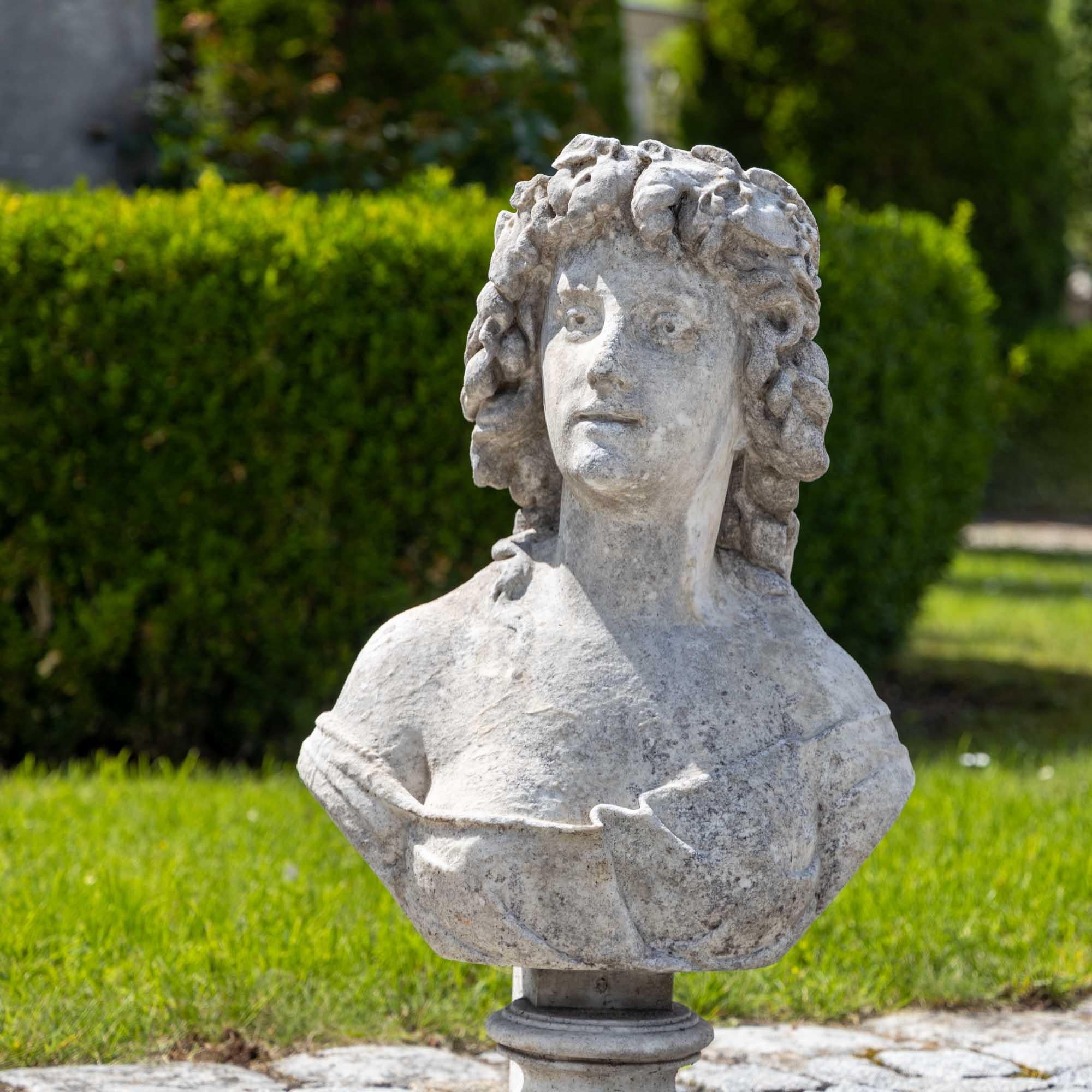 Hand carved marble bust of a woman with vine leaves in her curly hair and loosely falling clothes. The bust shows signs of weathering and has a slightly washed-out character.