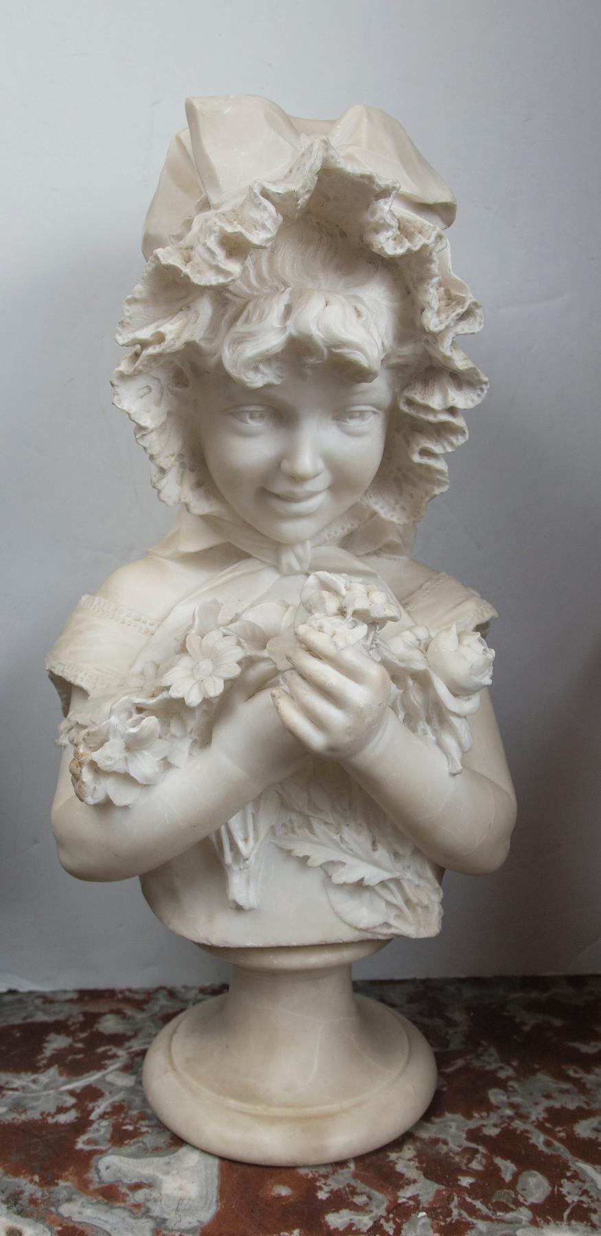 This half body marble bust depicts a female child holding a bunch of flowers to her chest. She wears a bonnet on her head, with front locks of hair showing. There is a bow, made of ribbon under her chin. There appears to be no signature.
Note the