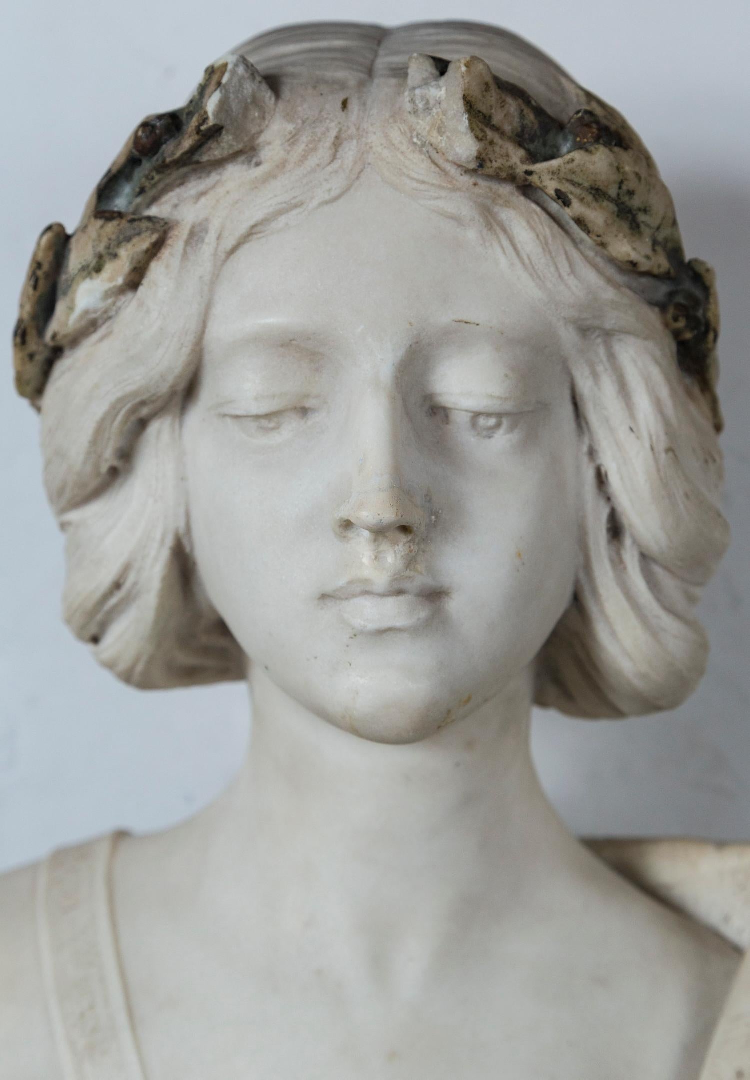 The white marble bust set upon a trapazoid shaped darker, veined marble. That part inscribed with the name SAPHO. She wears a laurel wreath atop her head. It has traces of paint, which may or not be original. The marble wreath is broken, with parts