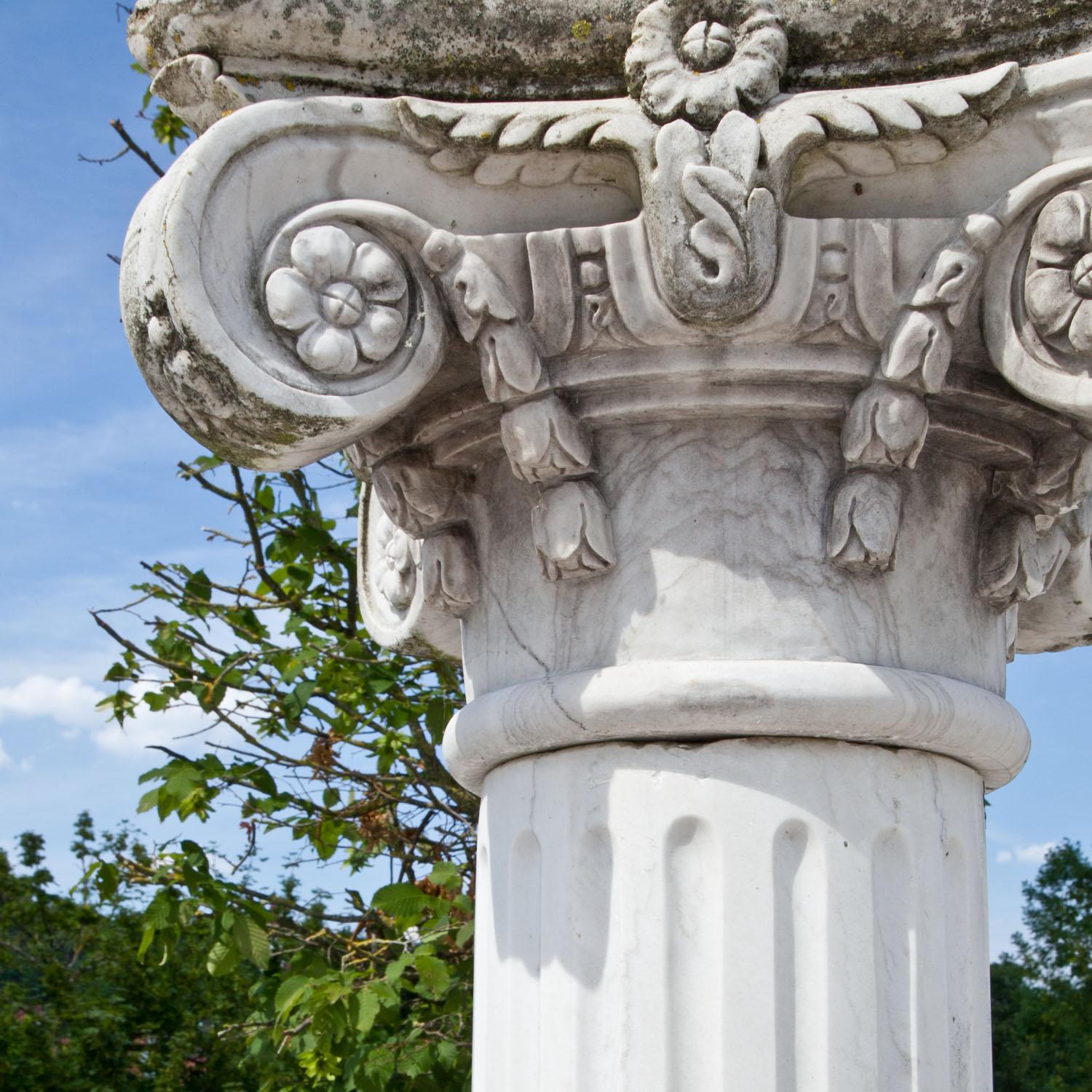 Grande colonne en marbre blanc et patine naturelle vieillie. La base est ronde, le fût est cannelé et le chapiteau est orné de volutes sculptées et de détails floraux.