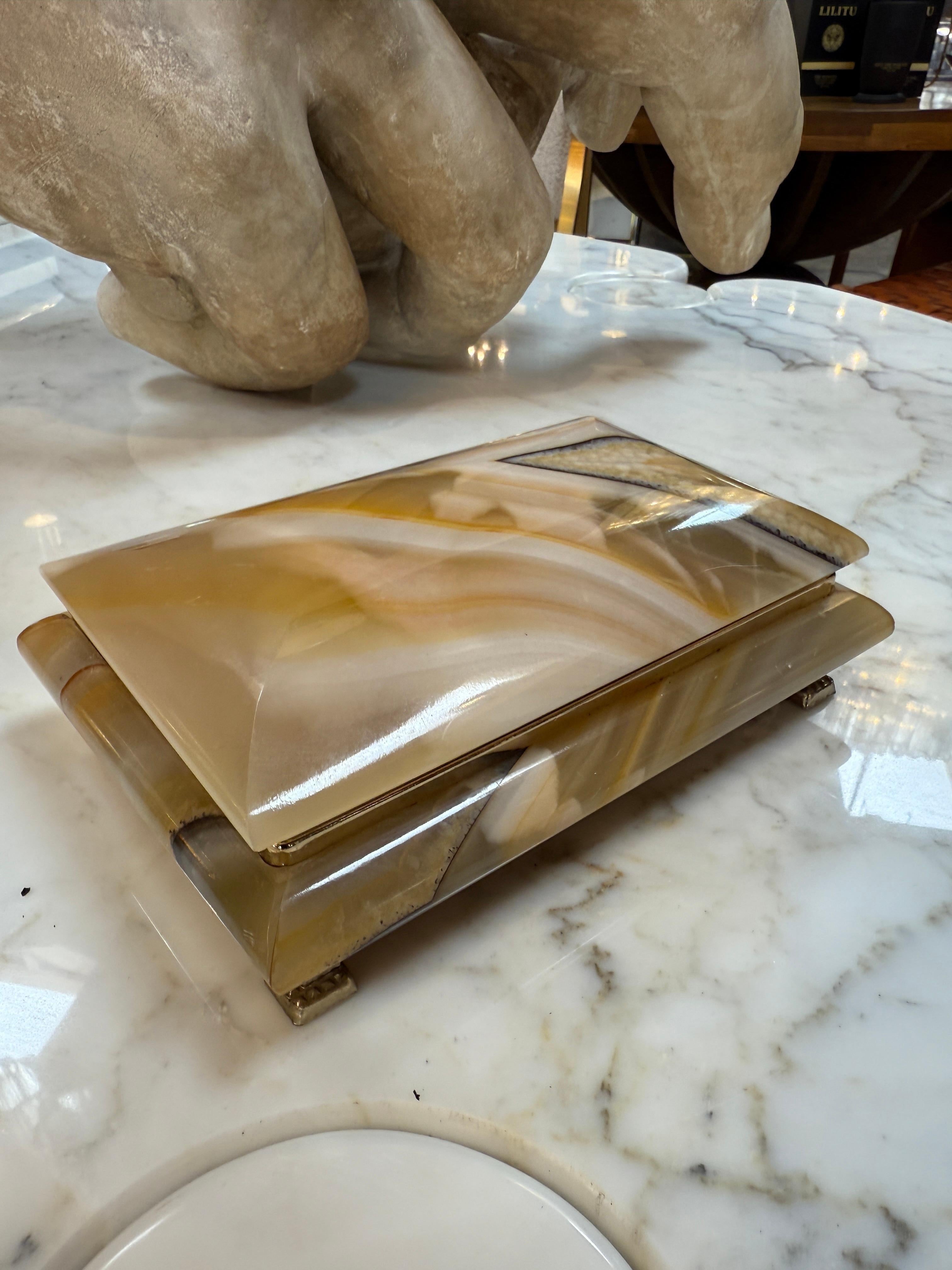 This richly detailed Italian decorative box showcases a polished onyx-style exterior with warm earth tones and distinctive natural veining. Resting on four small metal feet, it opens to reveal a vibrant red velvet-lined interior framed in brass.