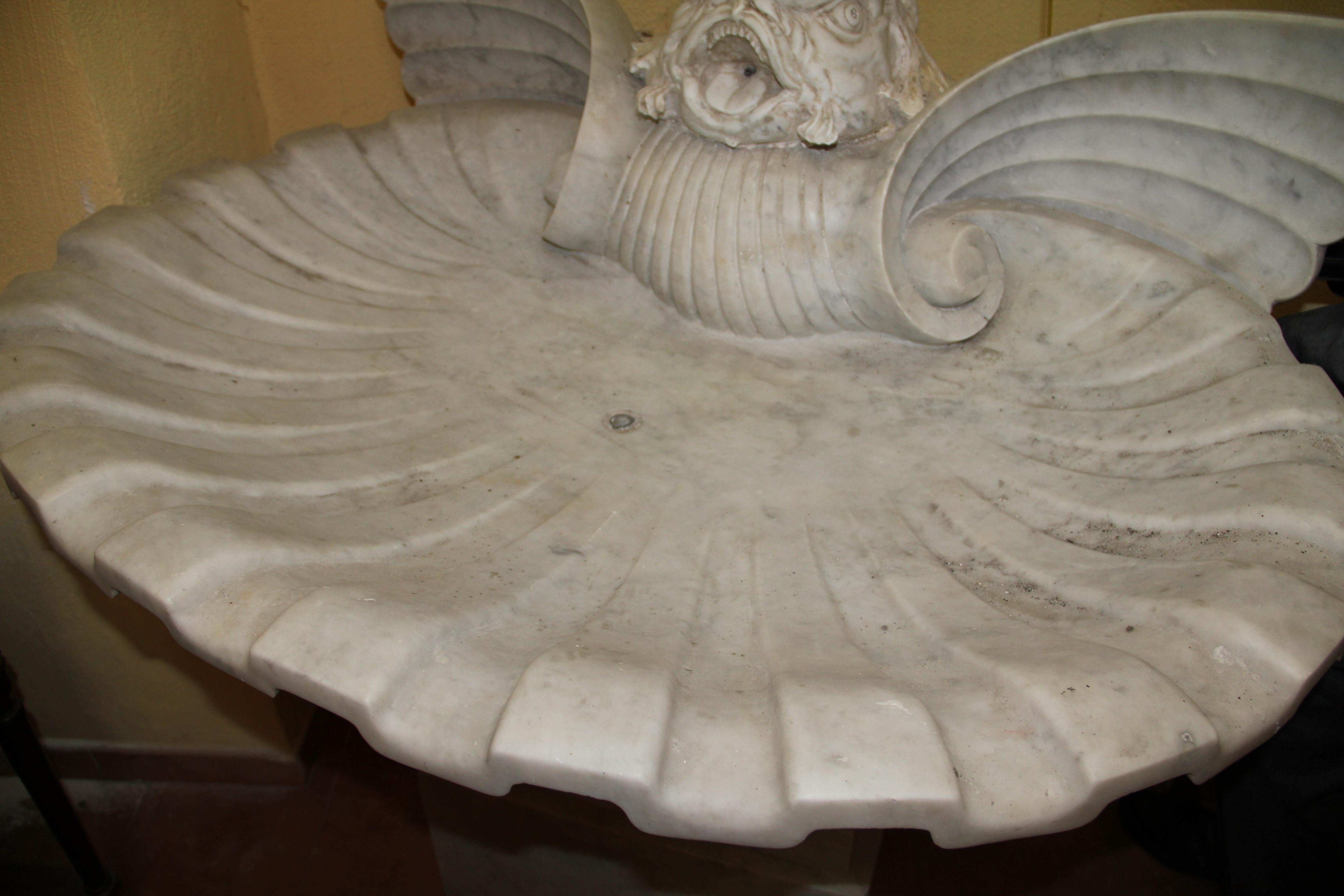 Carved of white marble, a stylised triton serving as spout, above a large scalloped sea shell, supported on organic, foliate scrolls.