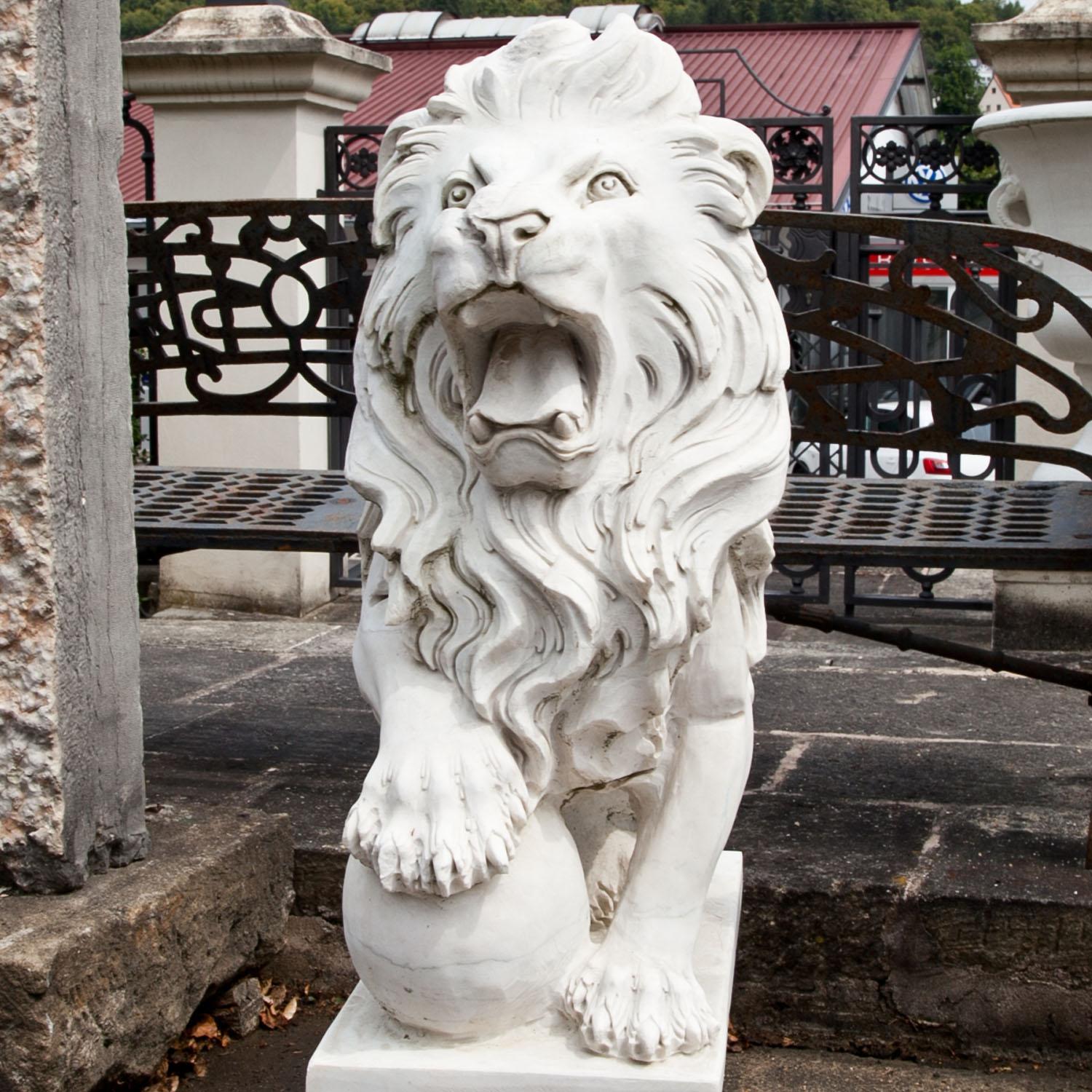 Pair of hand-carved male lions out of marble, standing on square bases. The lions are in a mirrored posture, each one placing his paw on a ball. They are depicted with an expressive mimic, wide-open mauls and a flowing mane.
