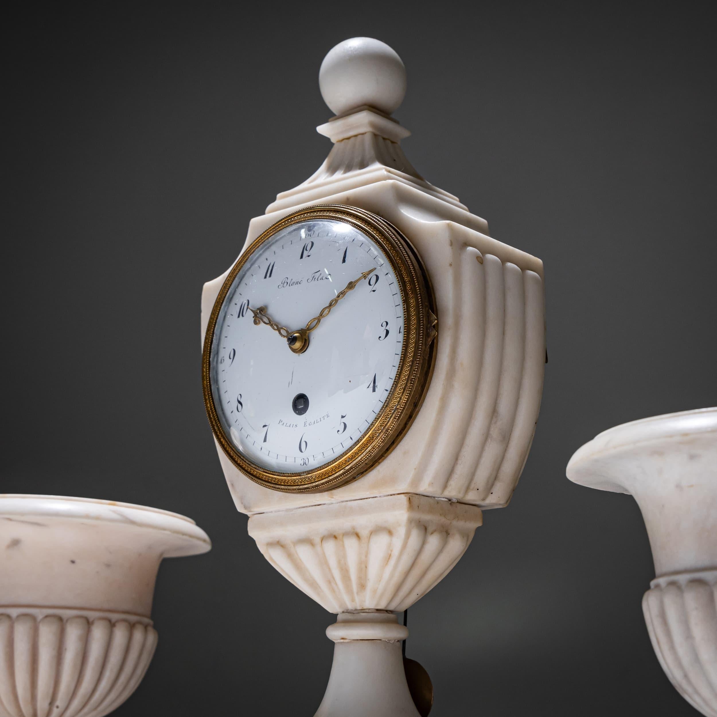 Refinado juego de guarnición neoclásico compuesto por un reloj de chimenea de mármol y dos jarrones a juego (20 x 13 x 13 cm). El reloj tiene un cuerpo en forma de jarrón de mármol blanco con detalles oscuros y una esfera redonda de esmalte rodeada