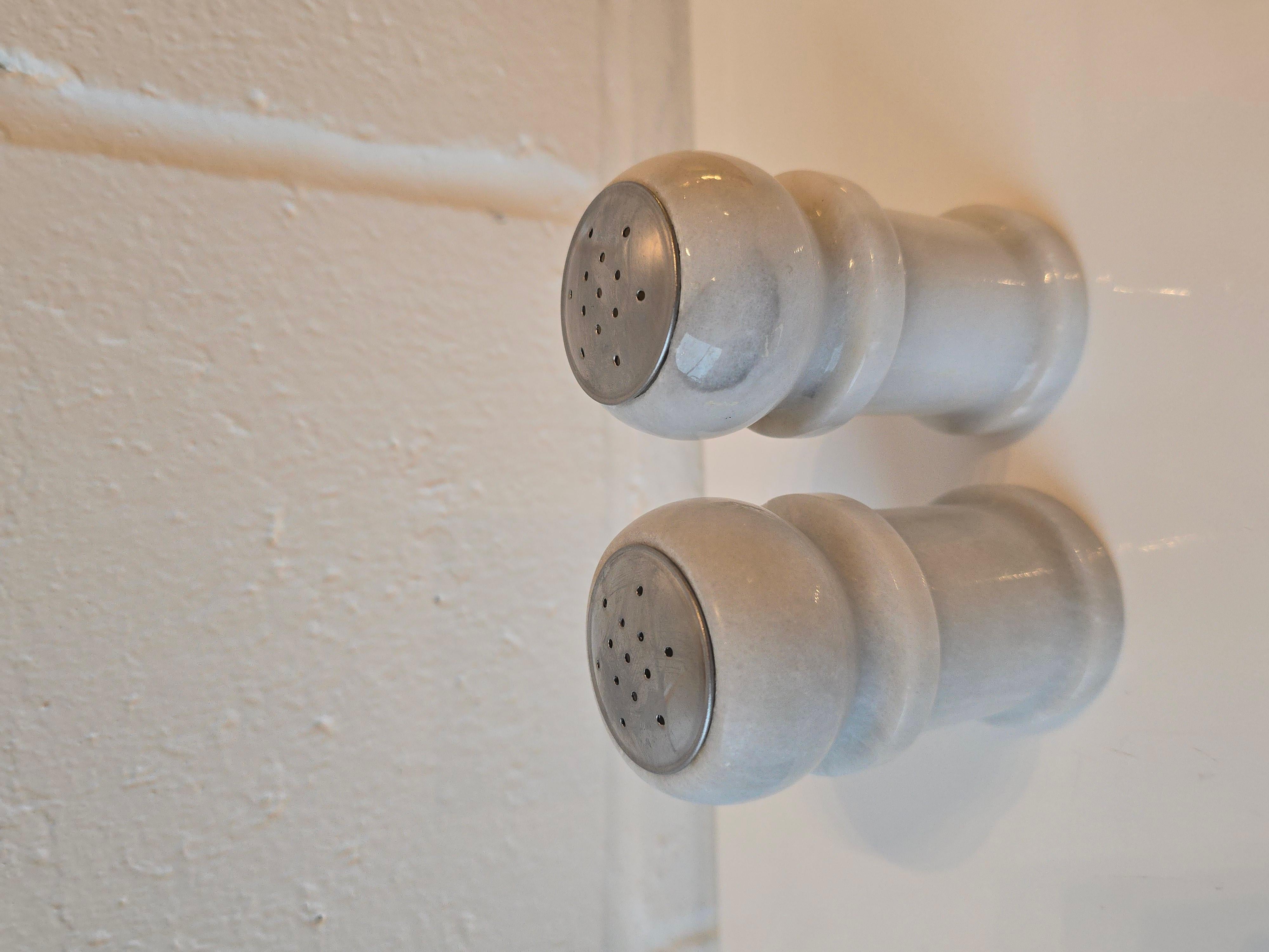 A pair of Carrera marble salt and peppers shakers. They are made out of carved Carrera marble, finished with a stainless steel top, a plastic cork at the bottom. While they are vintage, they appear to either be minimally used or unused. They have a