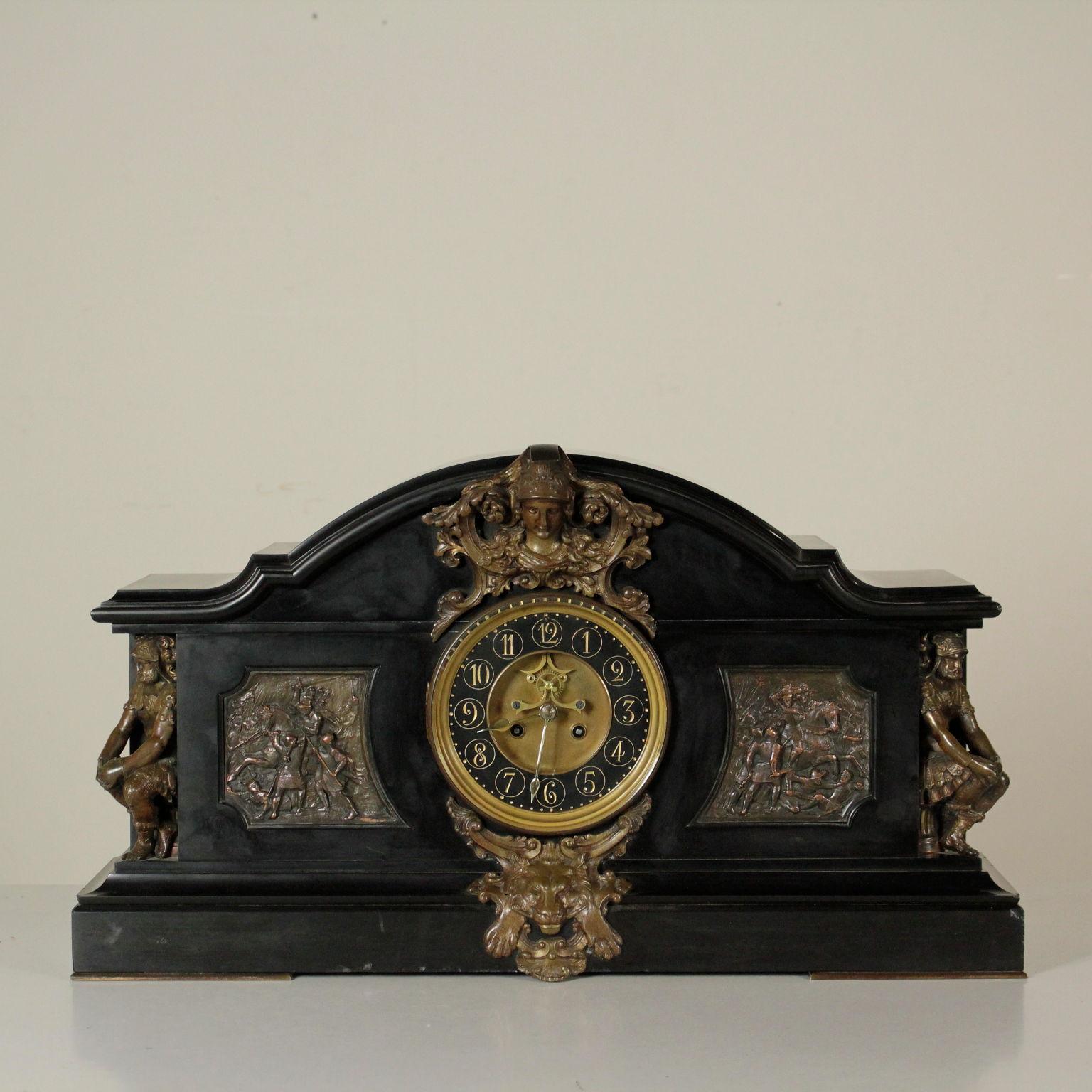 A table clock, black marble with bronze decorations. On the base, a frieze with lion head enclosed in a frame with volutes. On the sides, sculptures depicting soldiers seating on capitals. Battle scenes in low relief. Second dial made of bronze