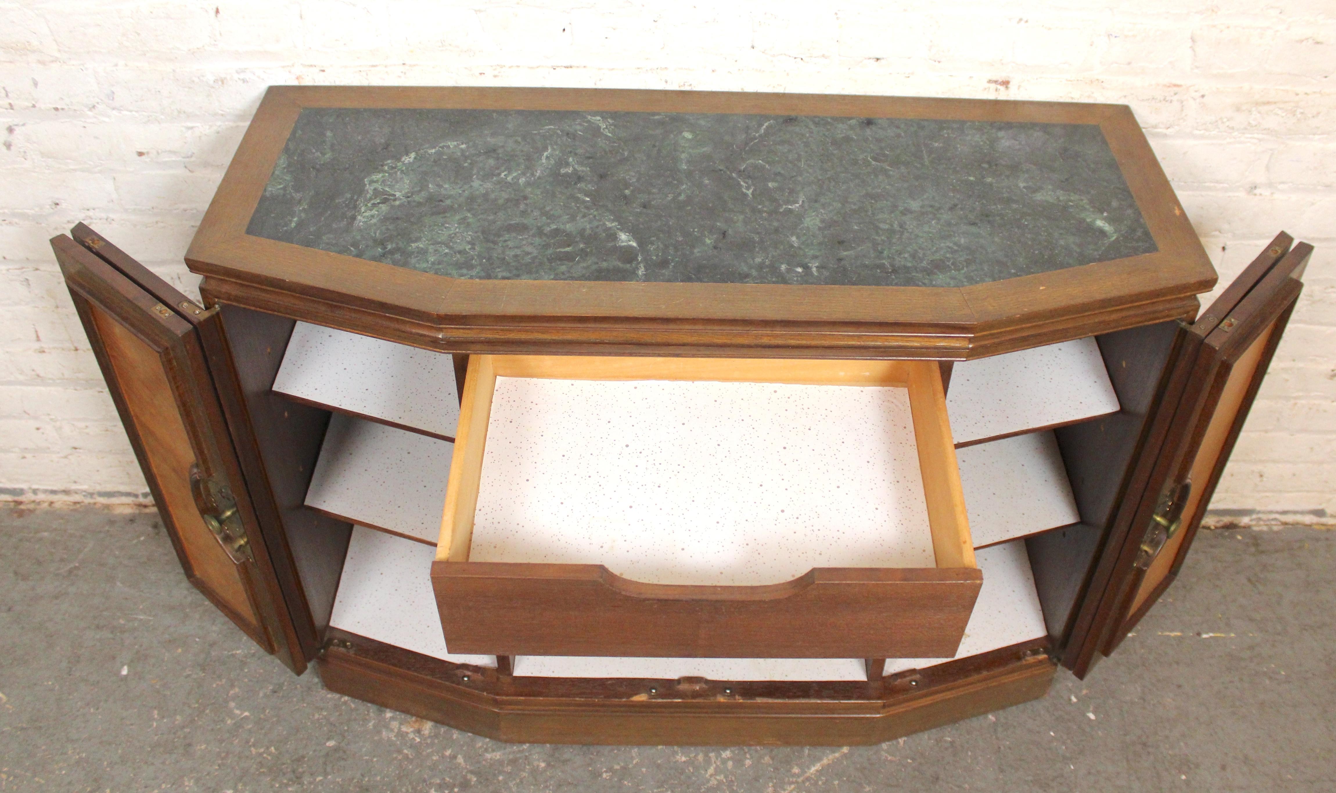 Cette console compacte présente une façade asiatique avec des plaques de laiton et des détails en bois sculpté. Deux portes Foldes s'ouvrent sur un grand espace de rangement avec des étagères réglables. Fini avec un plateau en marbre vert pour vos