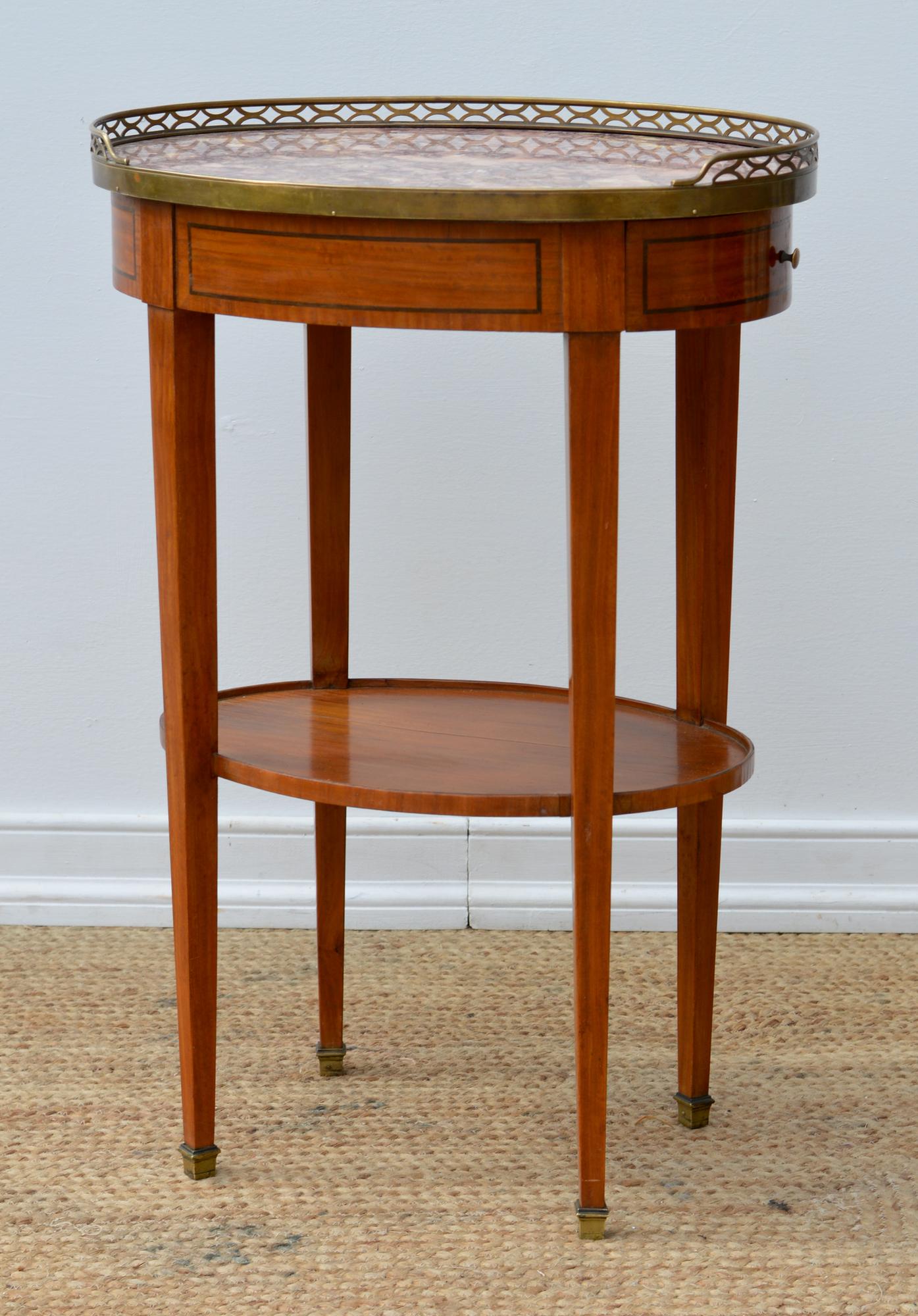 An attractive Directoire style tabourette marble topped side table having a pierced and wrapped brass gallery up top, and veneered and simply inlaid apron that supports one deep pulling drawer. A lower gallery of mahogany makes a useful spot for a