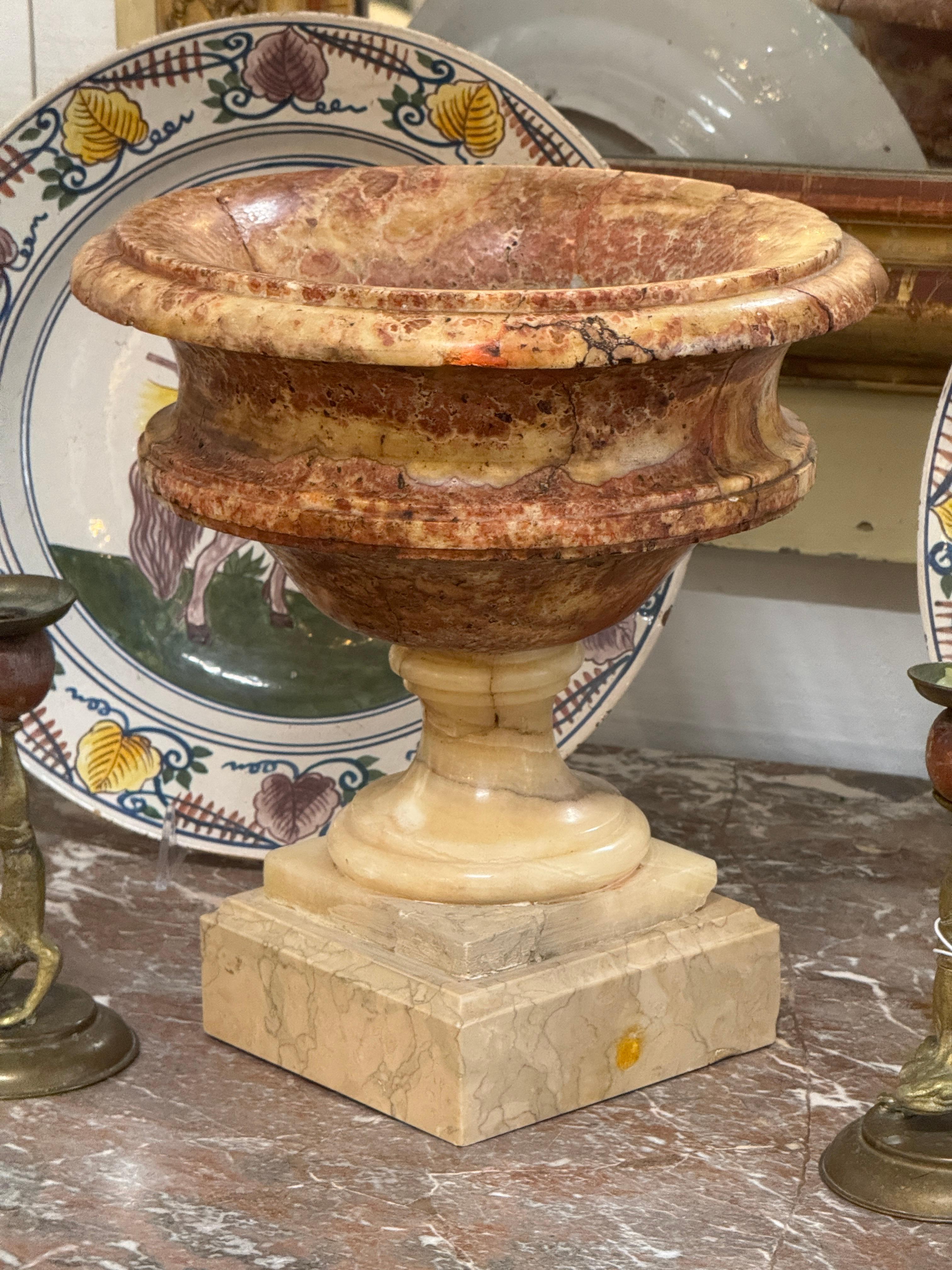 Marble urn with a flared rim and rounded body raised on a turned stem over a stepped square base. The stone shows varied veining and natural coloration throughout, with visible surface fissures typical of the material.