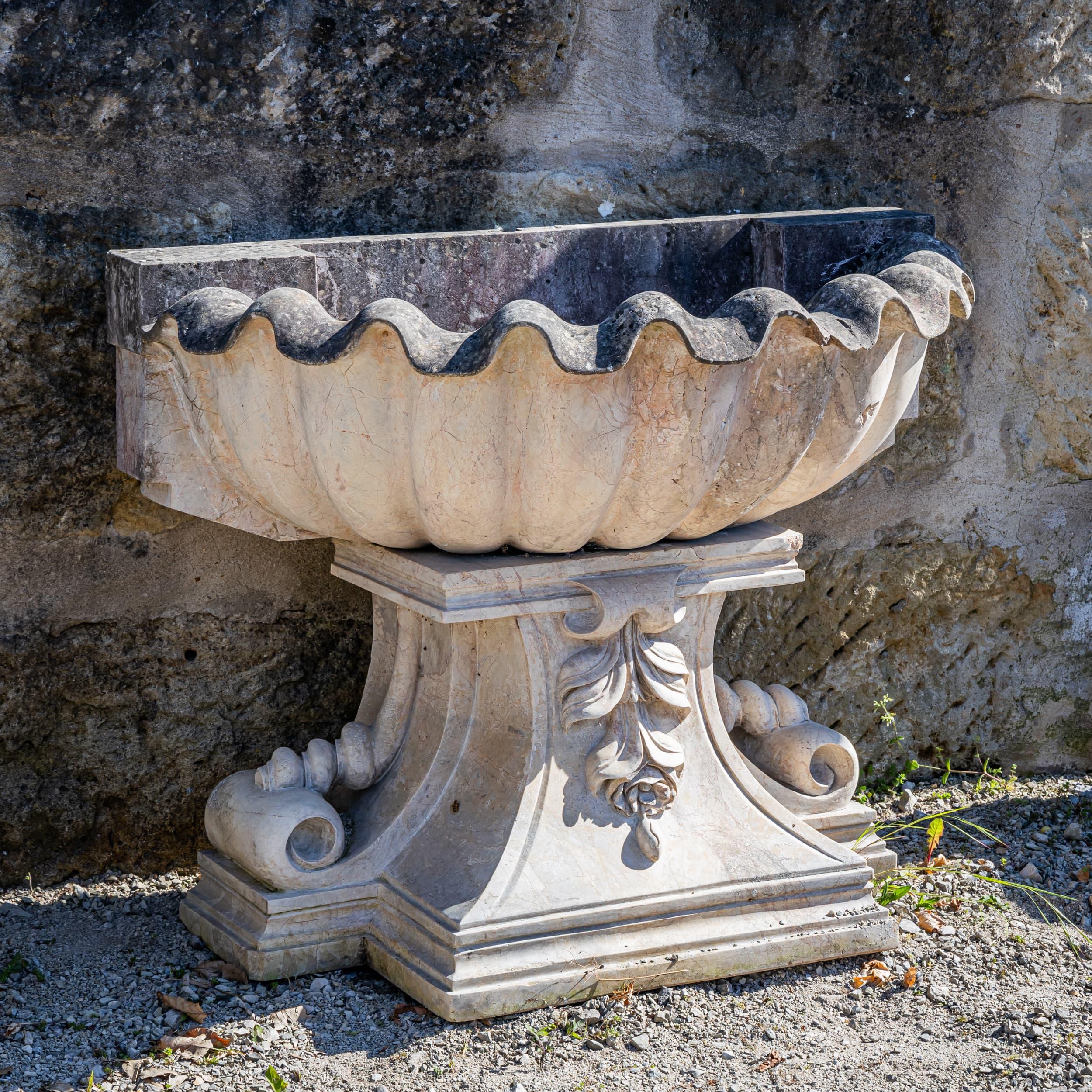 Fountain made of light marble with a shell-shaped bowl and richly decorated base. The finely sculpted volutes and the ornately carved leaf motif give the piece sculptural depth and classical elegance.