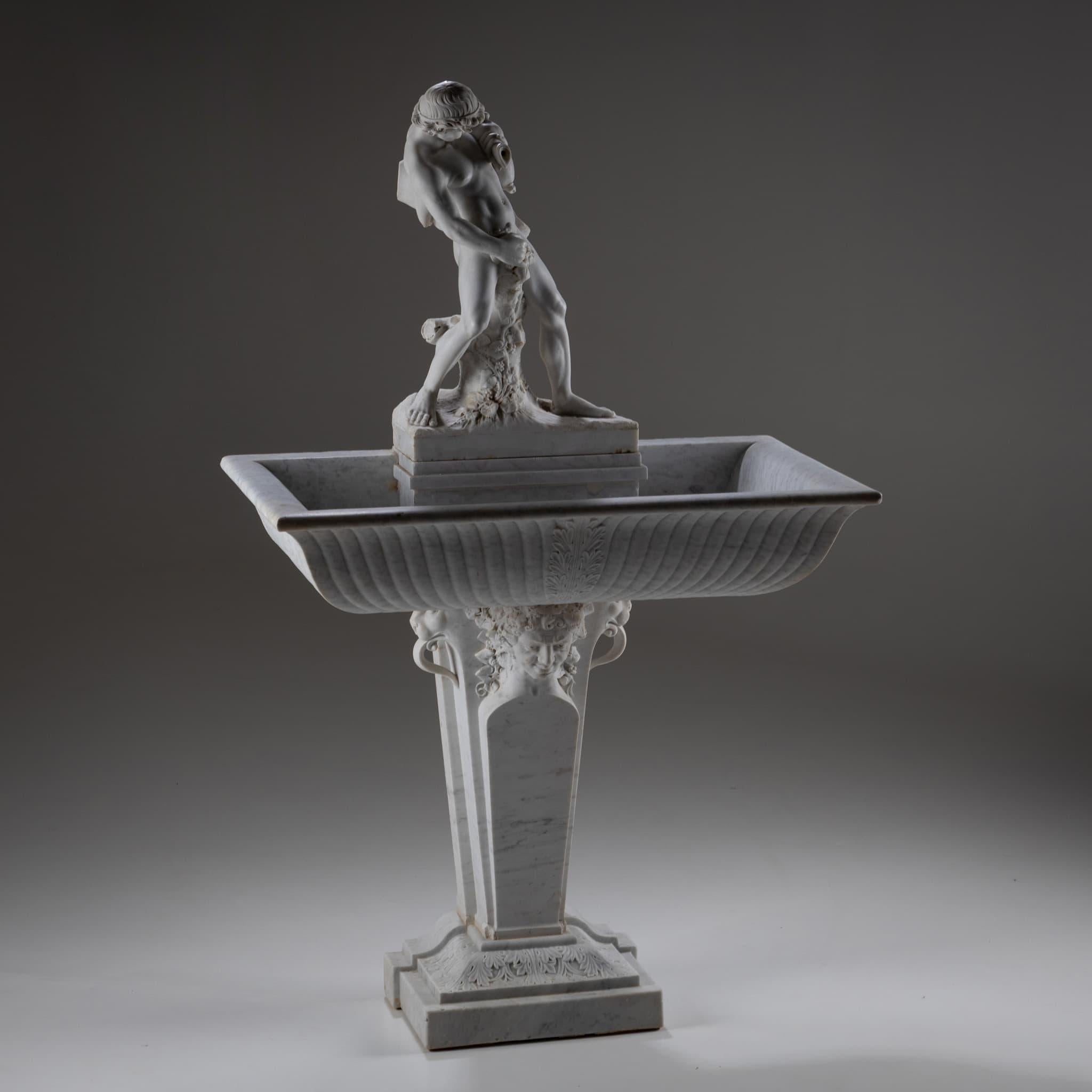 Trapezoidal marble wall fountain with a rectangular basin featuring gadrooned sides and leaf ornamentation. The basin rests on a pedestal adorned with a satyr protome, its hair entwined with vine leaves, flanked by lion masks with extended tongues.