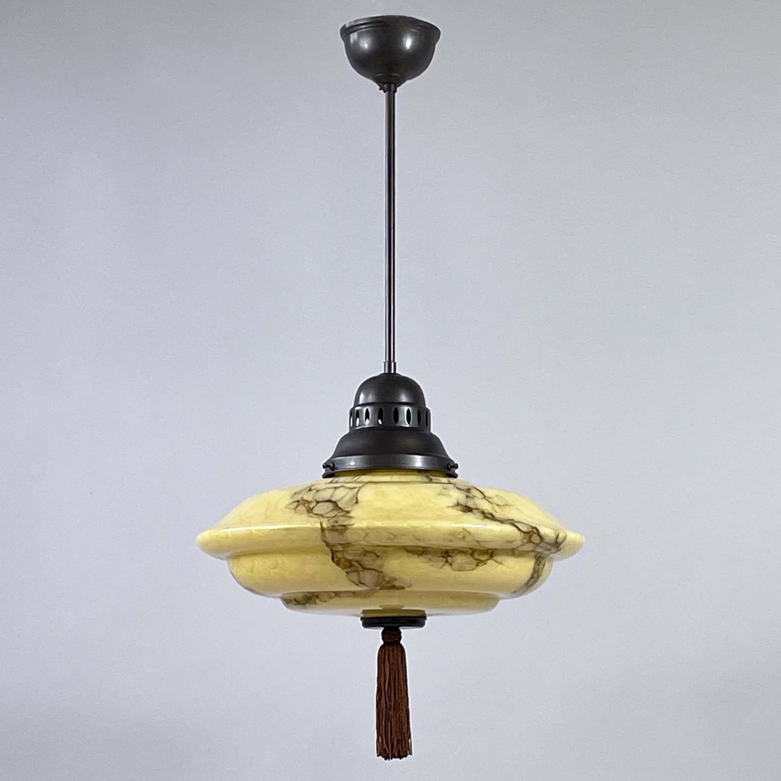 Este colgante Art Déco fue diseñado y fabricado en Alemania en la década de 1930.

La lámpara presenta una escultural pantalla de cristal jaspeado en cálidos tonos arena/amarillo con suaves vetas marrones, formando una silueta suave y estratificada