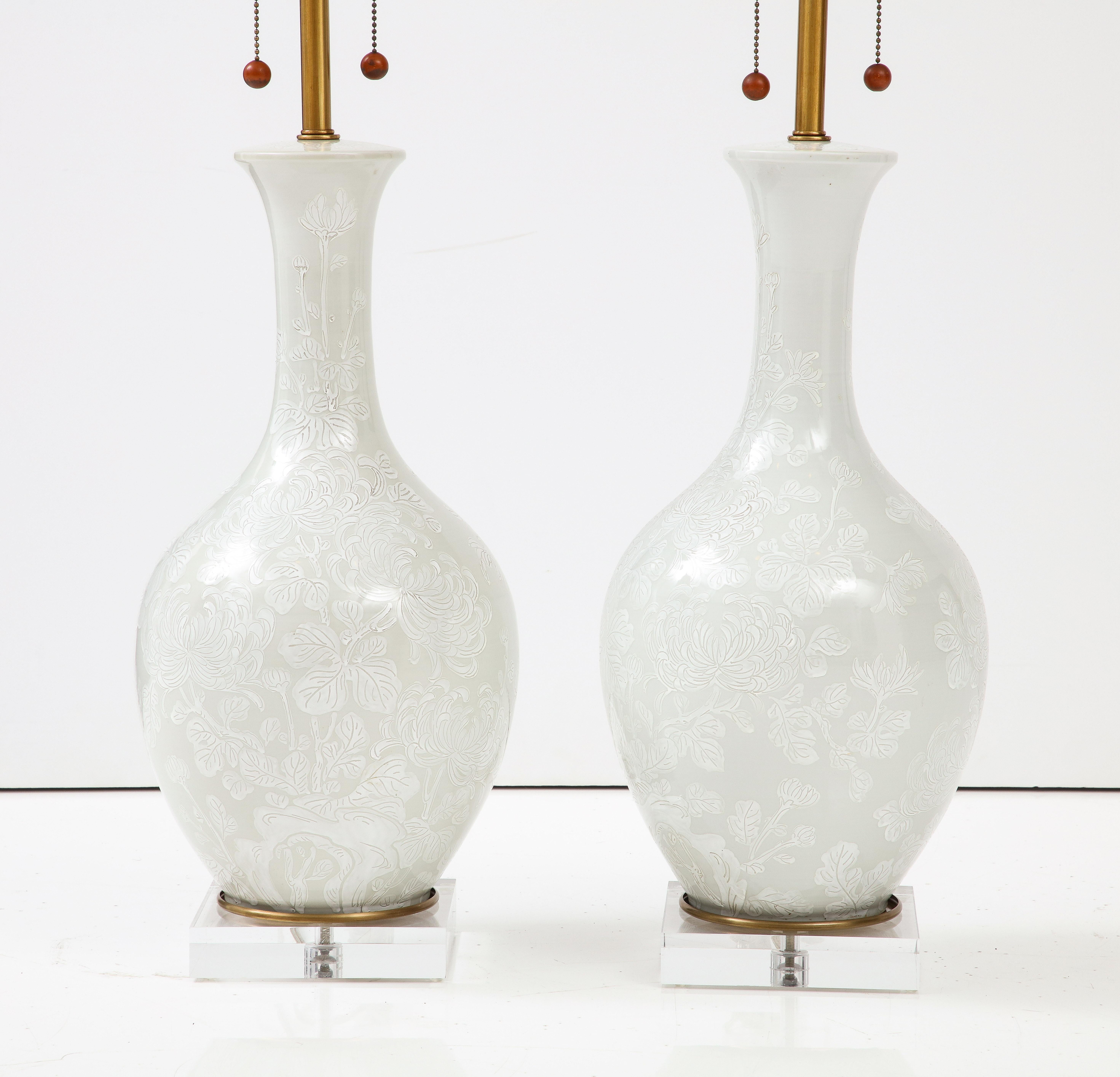 Mid Century porcelain lamps in an ivory white glaze embellished with white chrysanthemum pattern, resting on custom lucite bases. Double pull chains have been rewired, 100W bulb max. Marbro Lamp Co. 100W max bulbs.