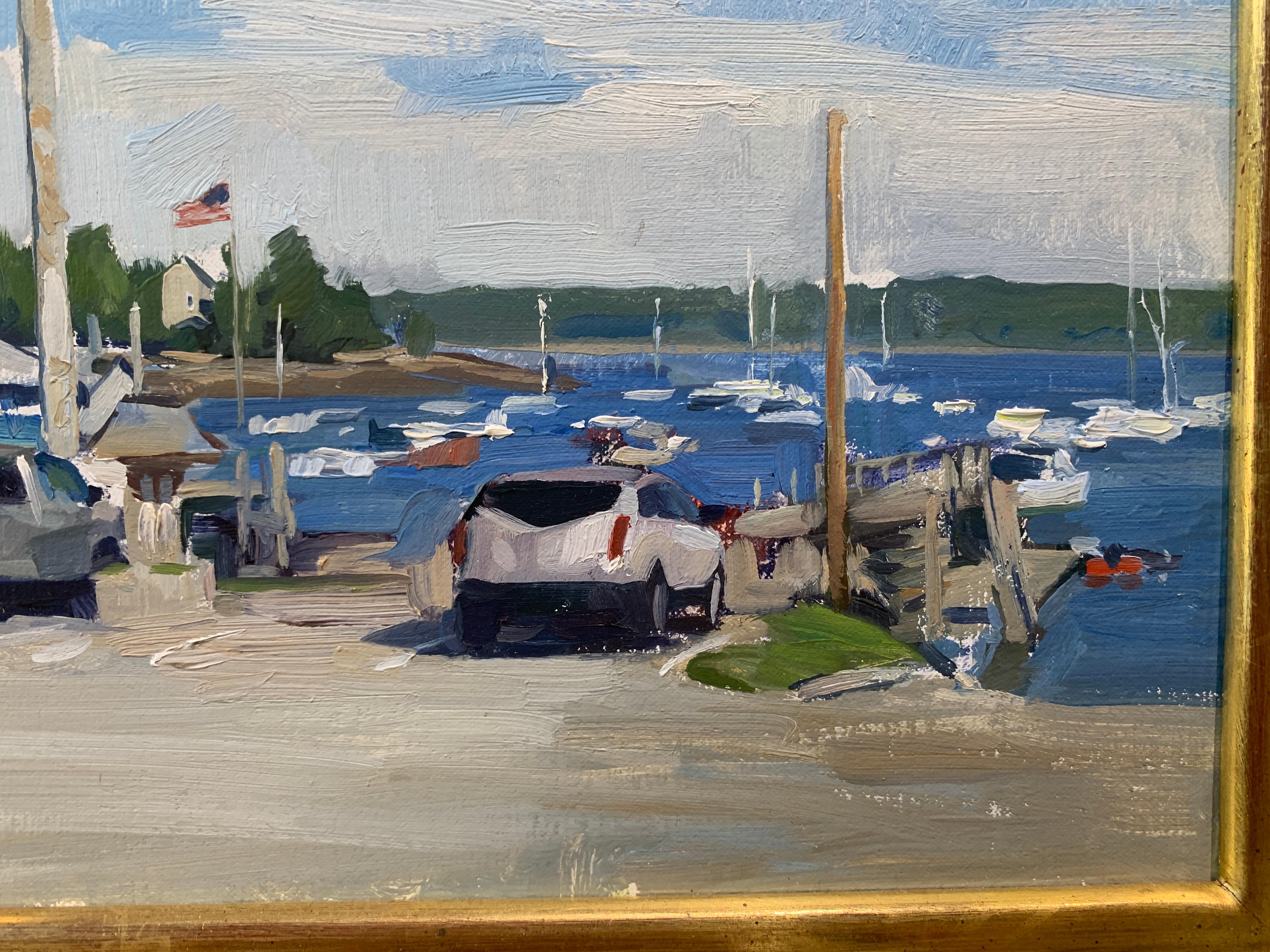 An oil painting, painted en plein air in the parking lot of the marina on Round Pond, Maine. Four cars are depicted parked along the entrance to the marina. An American flag can be seen flapping in the breeze. Boats sail in the pond in the distance.