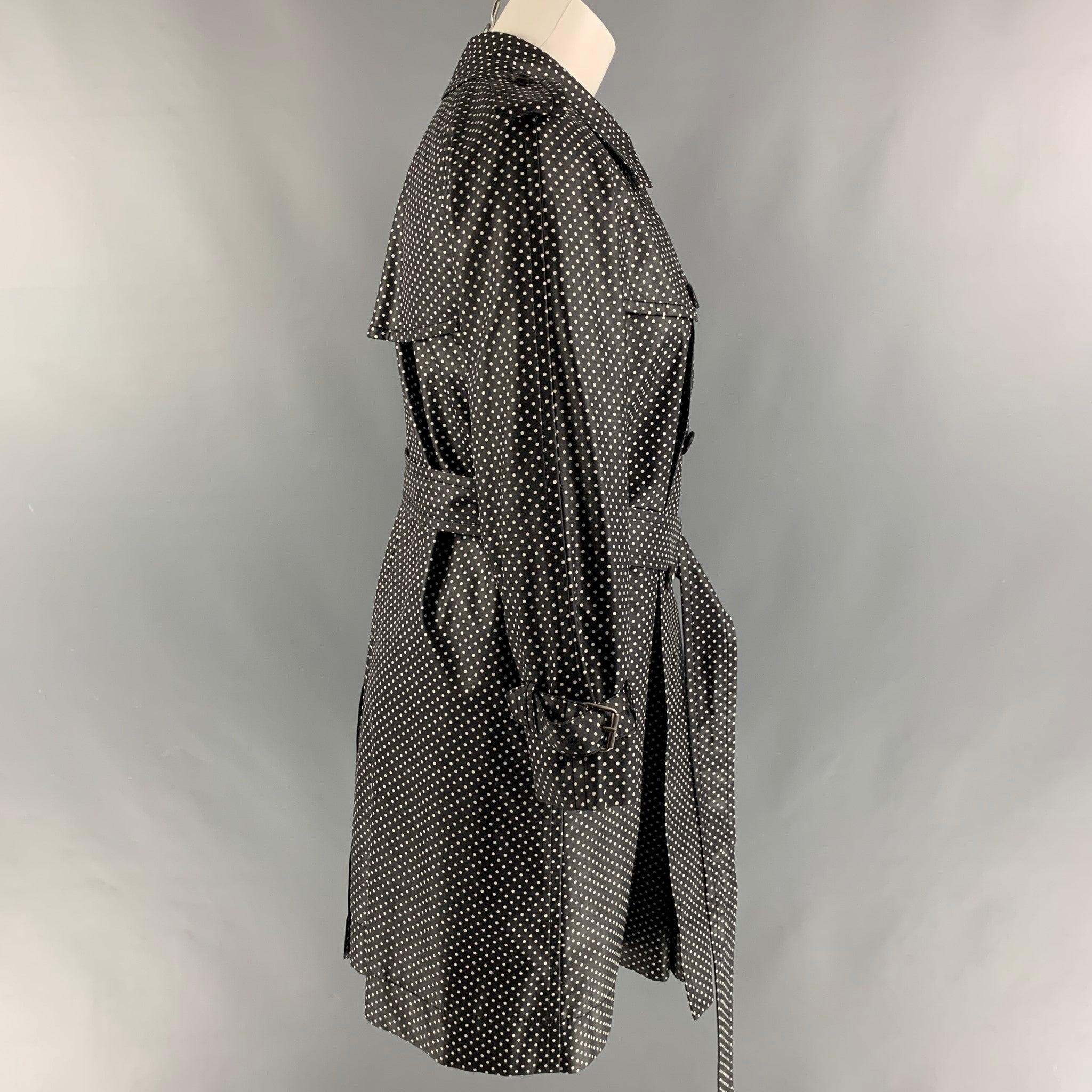 Le trench-coat MARC JACOBS est en soie à pois noirs et blancs, avec une doublure complète, un style ceinturé, une seule fente au dos, des épaulettes, une fermeture à crochet au col, des poches sur le devant et une fermeture à double boutonnage.