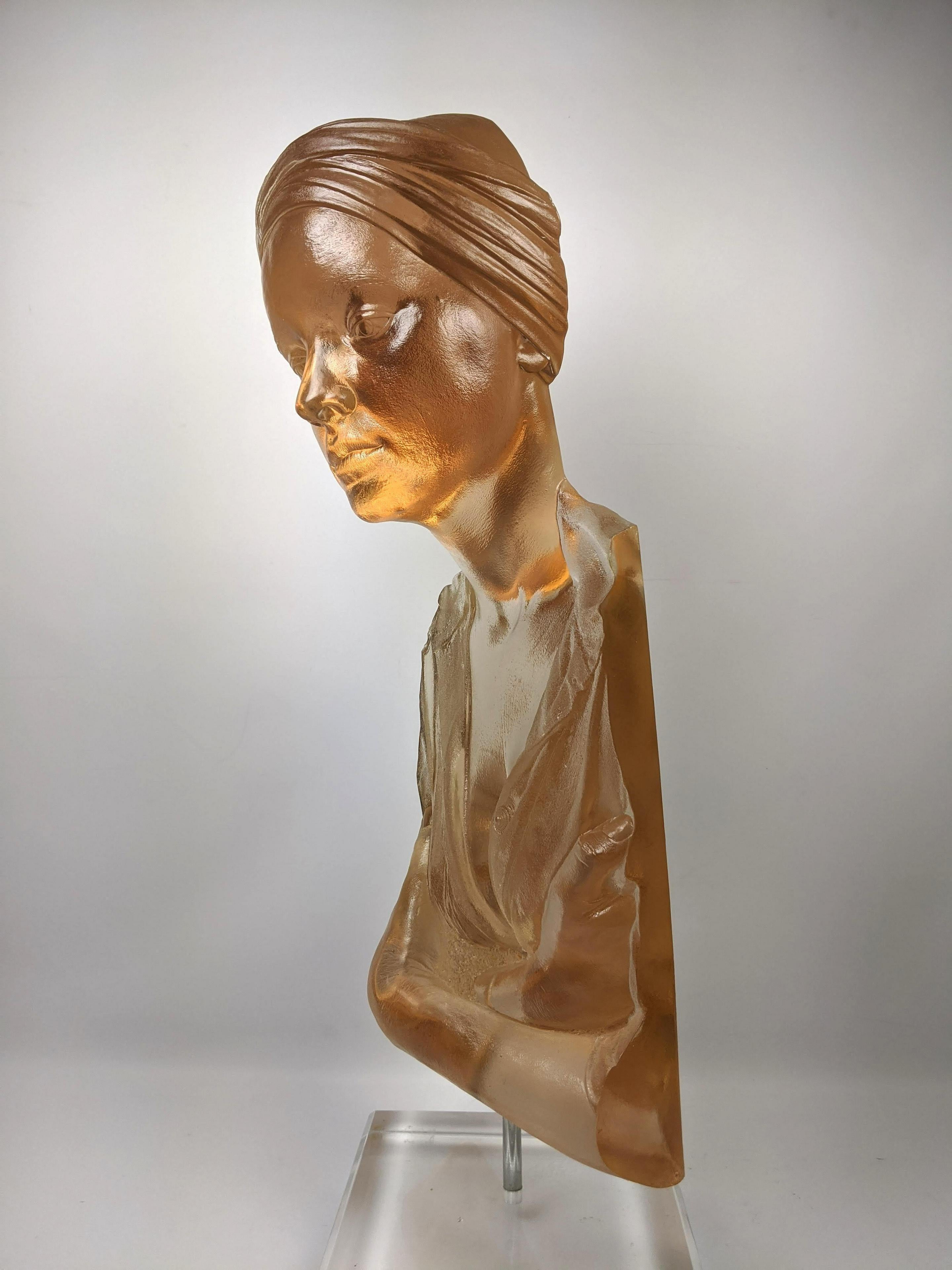 A cast acrylic sculpture titled Chin Up by American artist Marc Sijan. This sculpture is made from acrylic and portrays the upper torso of a clothed woman wearing a bandana over her free-flowing hair. Her eyes are closed and her head is tilted up as