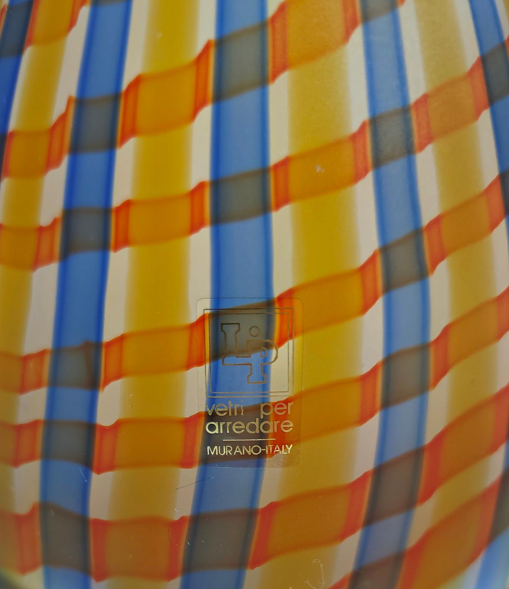 MARCELLO FURLAN. Labbro di produzione, Murano, Italia 1989 ca.
Vaso in vetro soffiato al latte decorato con canne intrecciate blu, arancione e giallo, corpo a clessidra con bocca circolare piatta. Label di produzione. Meravigliosa, la presenza ti