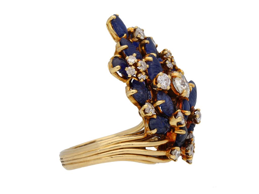 A yellow gold ring with heavy asymmetrical bezel composed of a drop shaped cluster of nineteen carved oval sapphires in claw settings with an approximate total weight of 20.00 carats, interspersed with two cushion shaped old mine diamonds in claw