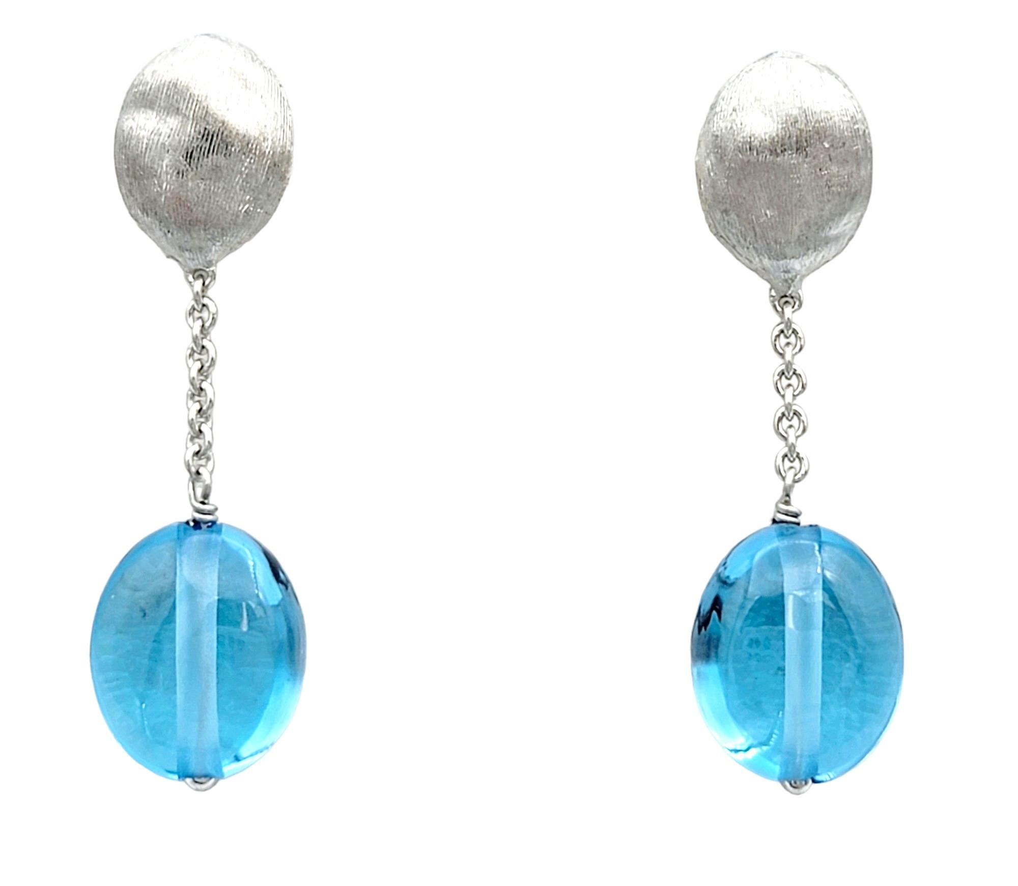 This gorgeous pair of Marco Bicego Siviglia blue topaz dangle earrings are a stunning display of elegance and sophistication, crafted with meticulous attention to detail in luxurious 18 karat white gold. Each earring features a captivating cabochon