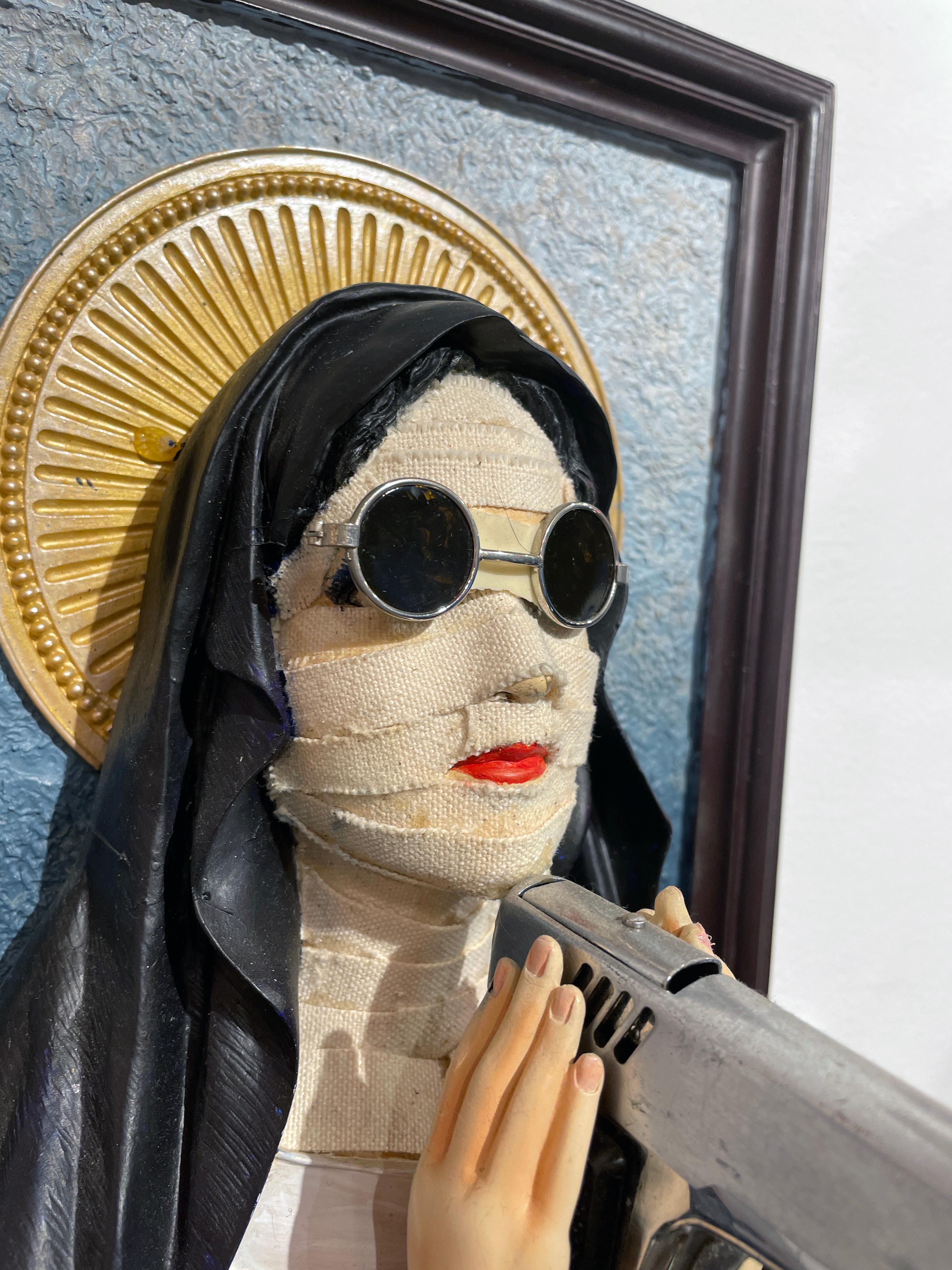 A found devotional sculpture of a woman in a habit becomes a caustic metaphor about the state of contemporary culture with a nun clasping a gun between her praying hands, sun glasses cover her eyes and her entire face shrowded in bandages.  Such is