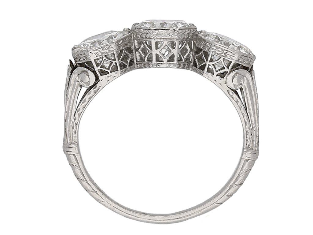 Three stone diamond ring. Horizontally set with three round old cut diamonds in open back grain set octagonal collets with an approximate combined weight of 3.13 carats, set to extremely ornate pierced open lattice collets with intricate pattern