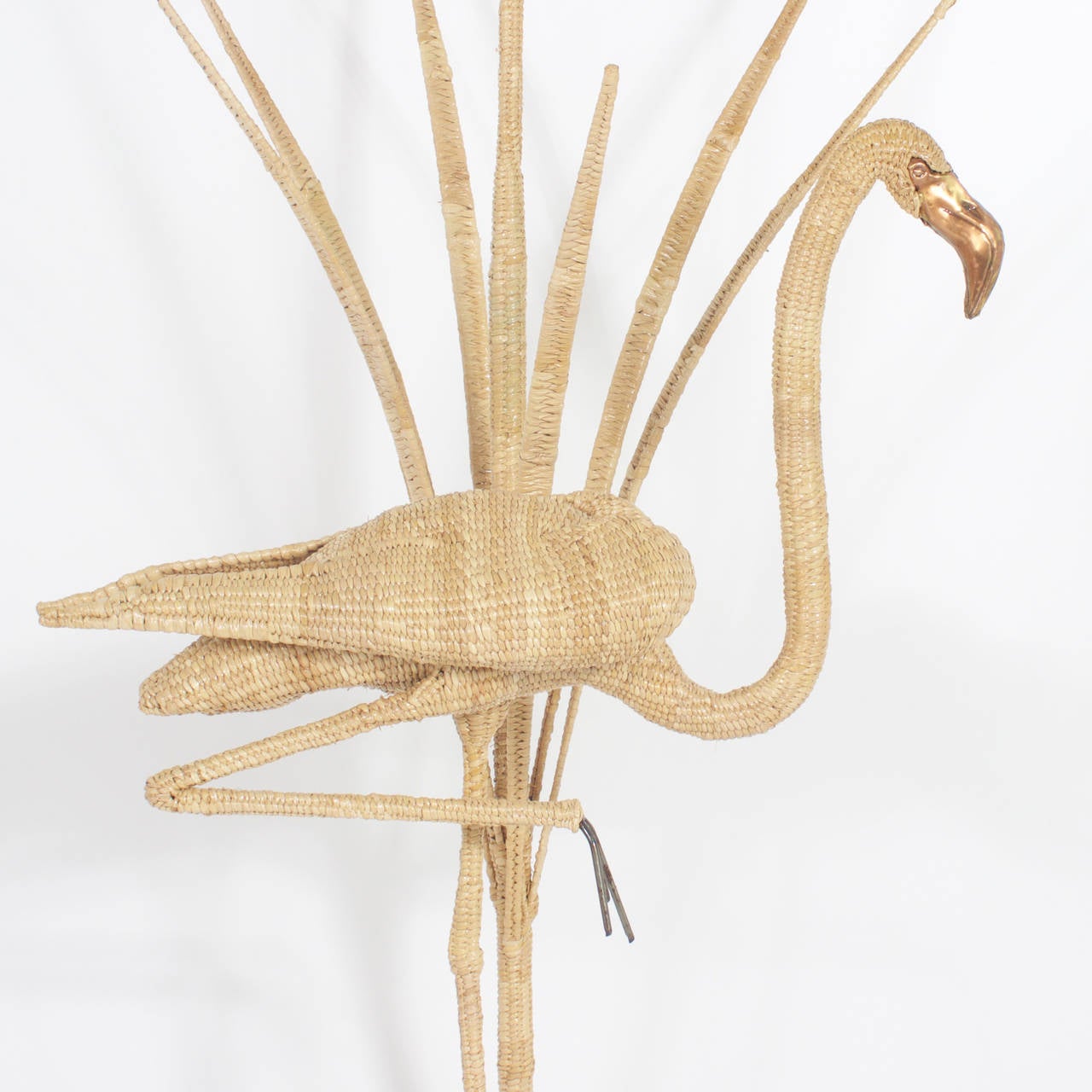 Mario Torres Flamingo floor lamp striking a familiar one legged pose. Constructed of a metal frame wrapped in wicker or reed. Featuring brass beaks and eyes, standing in front of a patch of reeds. Signed Mario Torres, EST. 1974 on a brass medallion.