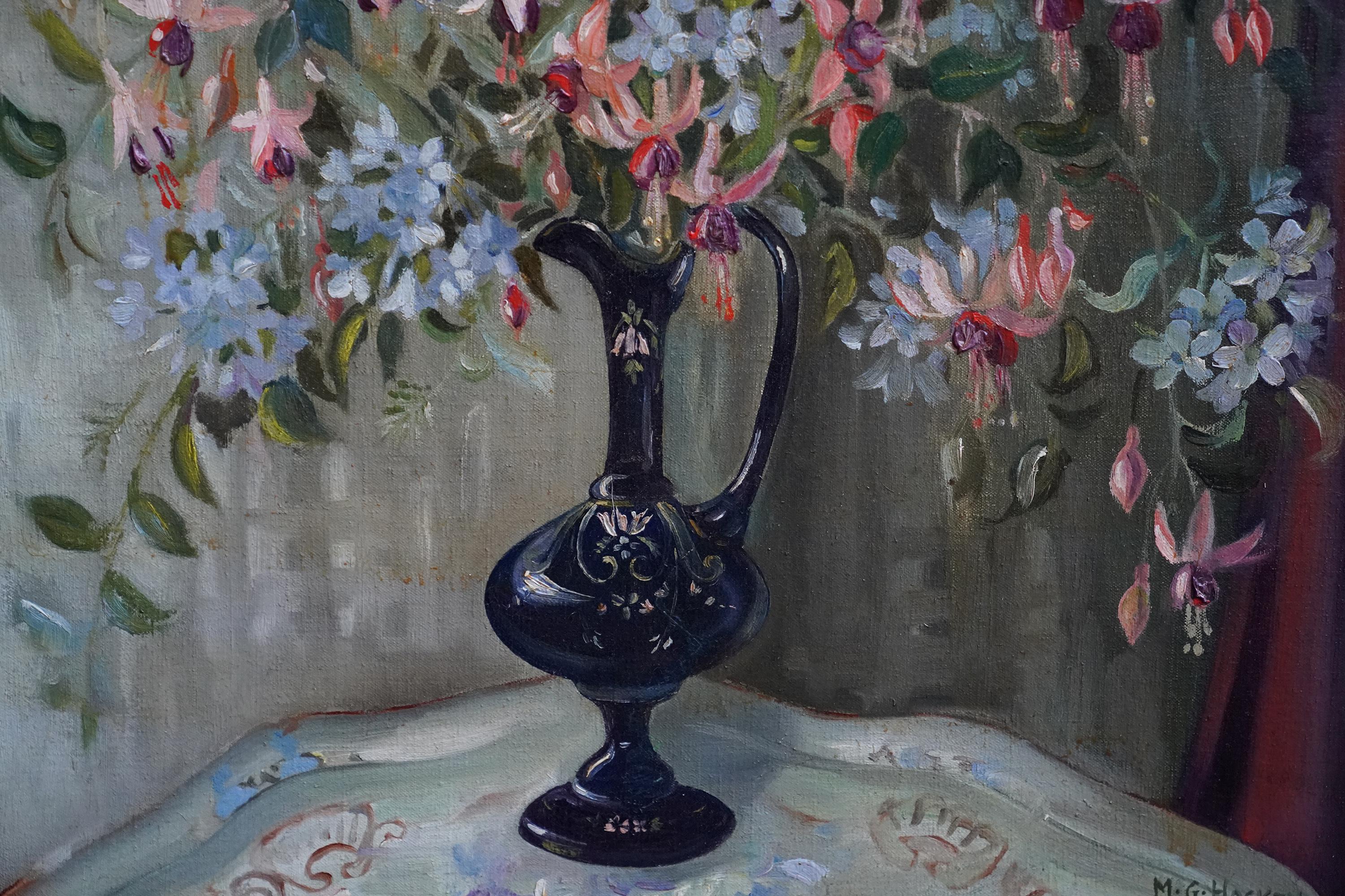 This charming floral oil painting is by Cornish born artist Marion Grace Hocken. Painted circa 1950 it is a floral arrangement of pink and purple fuchsia with sprays of blue flowers in a blue glass vase on a table. A keen botanist, Hocken has