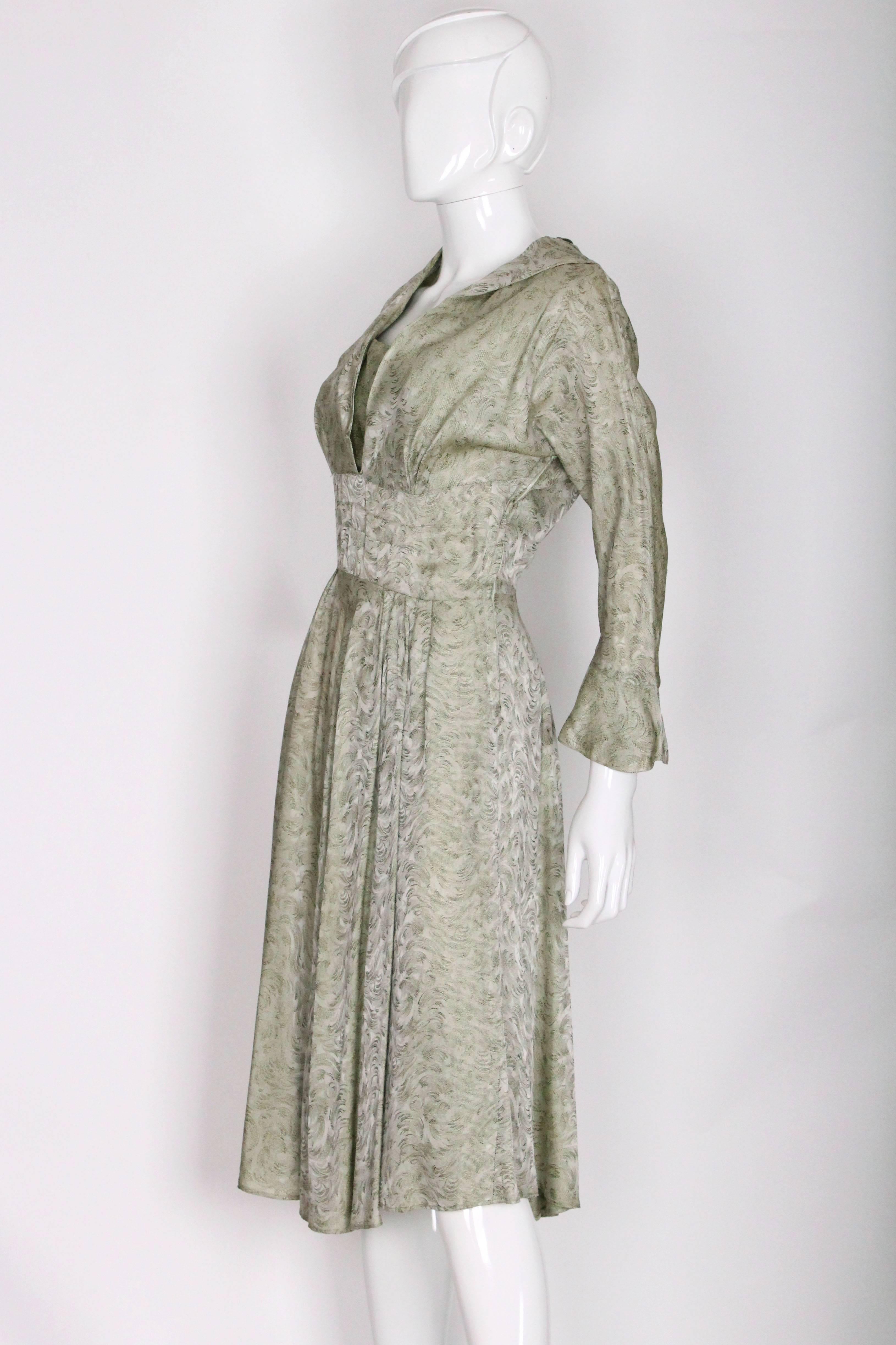 A cute day dress by Marion Michaels. In a light sage green jacquard and silk fabric with swirling design in tonal shades. This dress has a chic frilled collar and cuffs and a fullish skirt that is gathered from the waist band. There is a small side