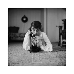 Audrey Hepburn con blusa blanca y teléfono, tumbada, con la mano en el receptor y la barbilla hacia abajo