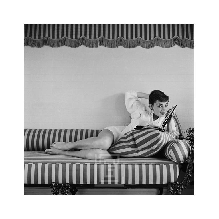 Black and White Photograph Mark Shaw - Audrey Hepburn sur canapé rayé, dossier, tête inclinée, soufflant debout, 1954