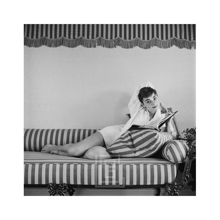 Black and White Photograph Mark Shaw - Audrey Hepburn sur canapé rayé, nœud derrière la tête, 1954