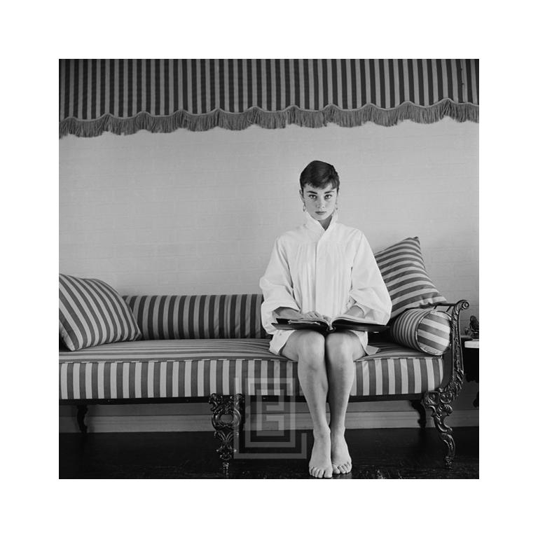 Mark Shaw Portrait Photograph - Audrey Hepburn on Striped Sofa, Faces Forward with Book Open, 1954