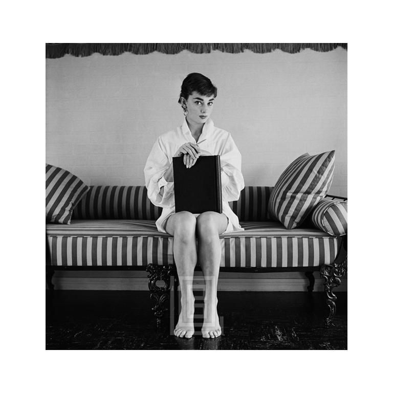 Black and White Photograph Mark Shaw - Audrey Hepburn sur canapé rayé, mains sur livre fermé, 1954