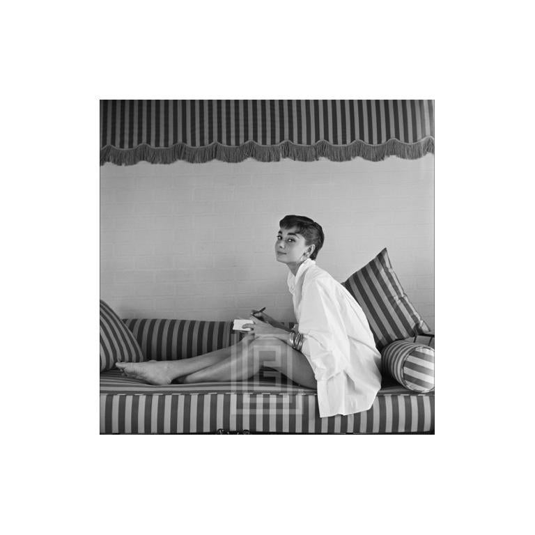 Mark Shaw Portrait Photograph - Audrey Hepburn on Striped Sofa, Leans Forward Writing, 1954