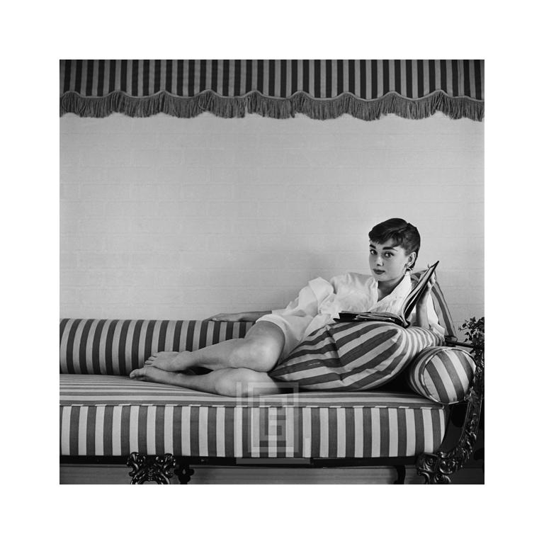 Mark Shaw Portrait Photograph - Audrey Hepburn on Striped Sofa, Reclines, Book Open, 1954