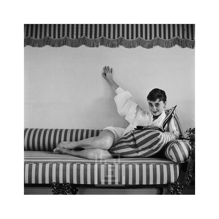 Mark Shaw Portrait Photograph - Audrey Hepburn on Striped Sofa, Reclines Hand Up, Book Open, 1954