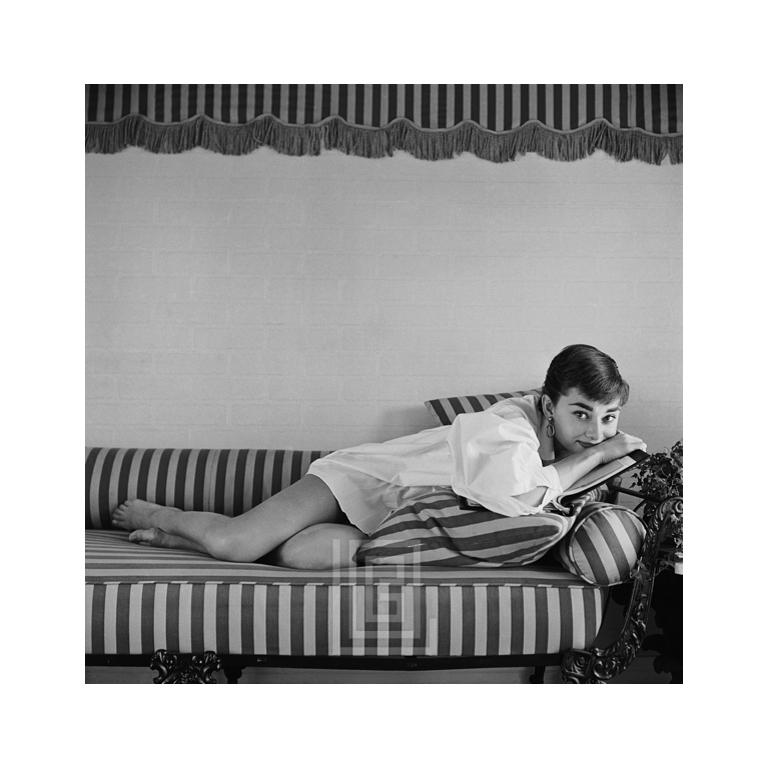 Black and White Photograph Mark Shaw - Audrey Hepburn sur canapé rayé, reposant sur un livre, 1954