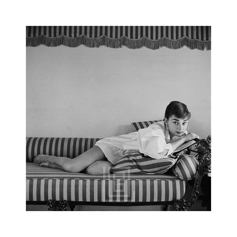 Black and White Photograph Mark Shaw - Audrey Hepburn sur canapé rayé, reposant sur une tête de livre, 1954