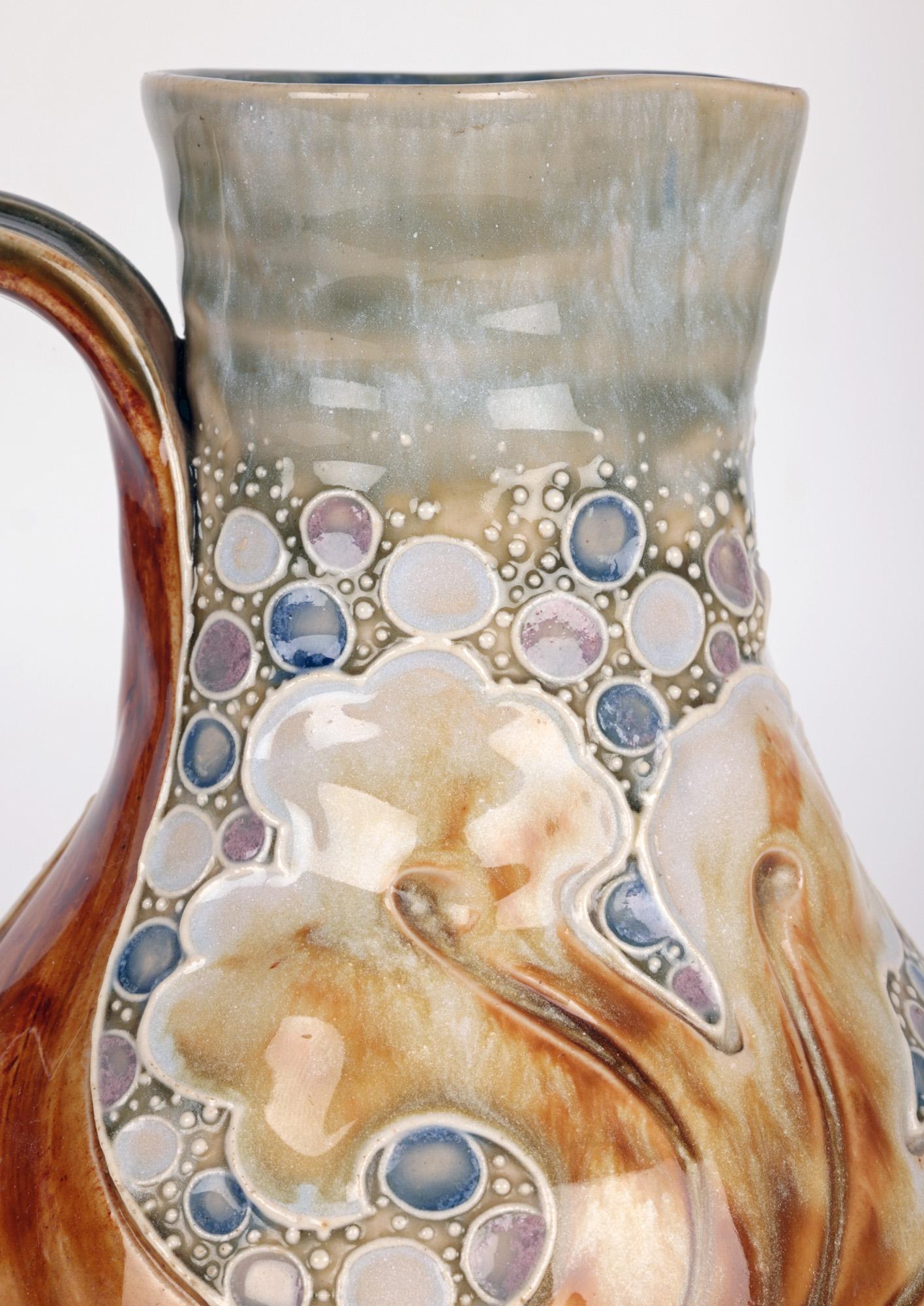 A very stylish and unusual Doulton Lambeth Art Pottery jug with mask handle and scrolling leaf designs by renowned modeler, designer and decorator Mark V Marshall and dating from around 1890. The stoneware jug is of rounded bulbous shape standing on