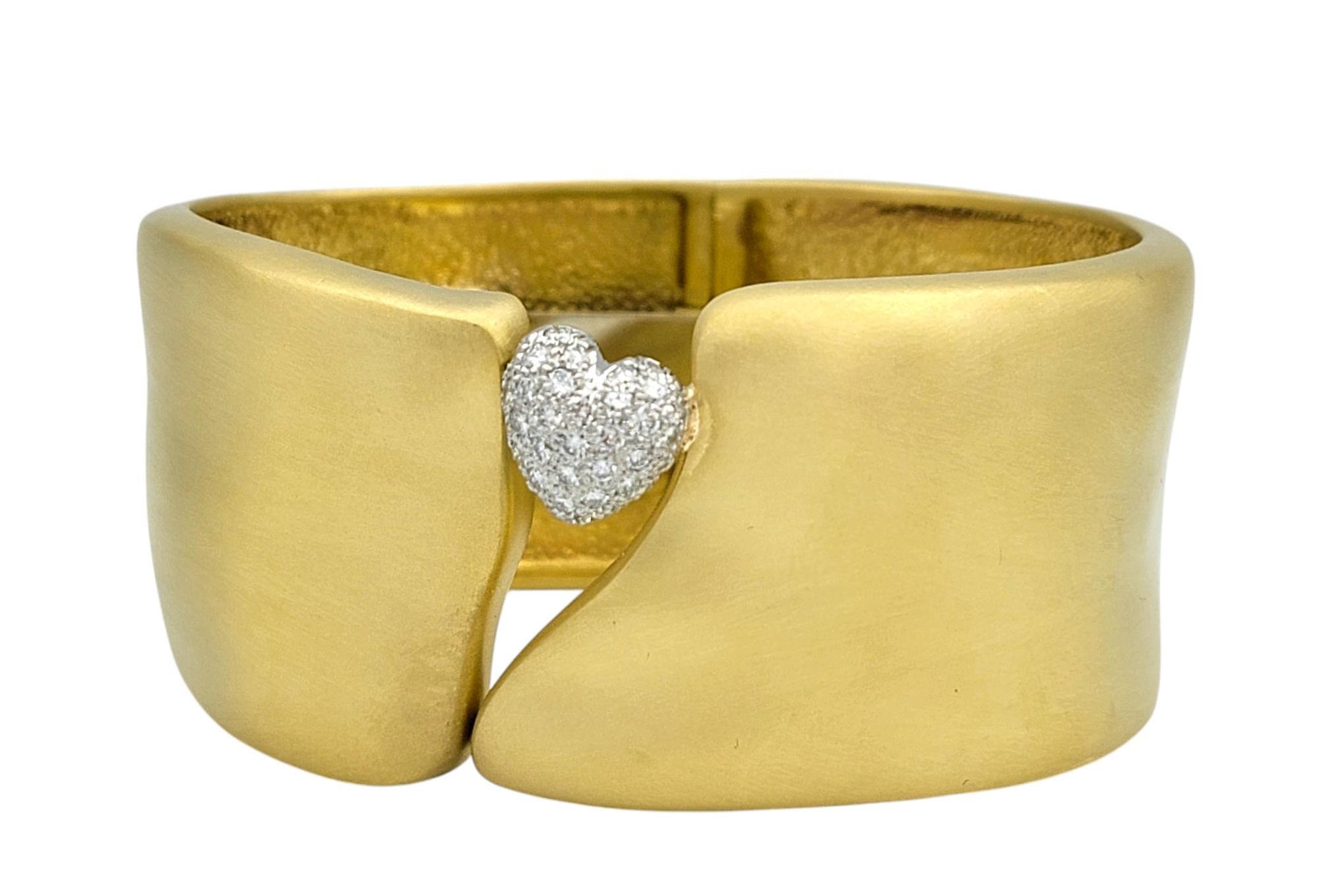 Dieses kühne, zeitgenössische Accessoire verleiht dem Handgelenk schlichte Eleganz. Das unglaubliche Manschettenarmband hat ein gebürstetes Gold-Finish, das von einem einzelnen glitzernden, gepflasterten Diamantenherz akzentuiert wird. Das