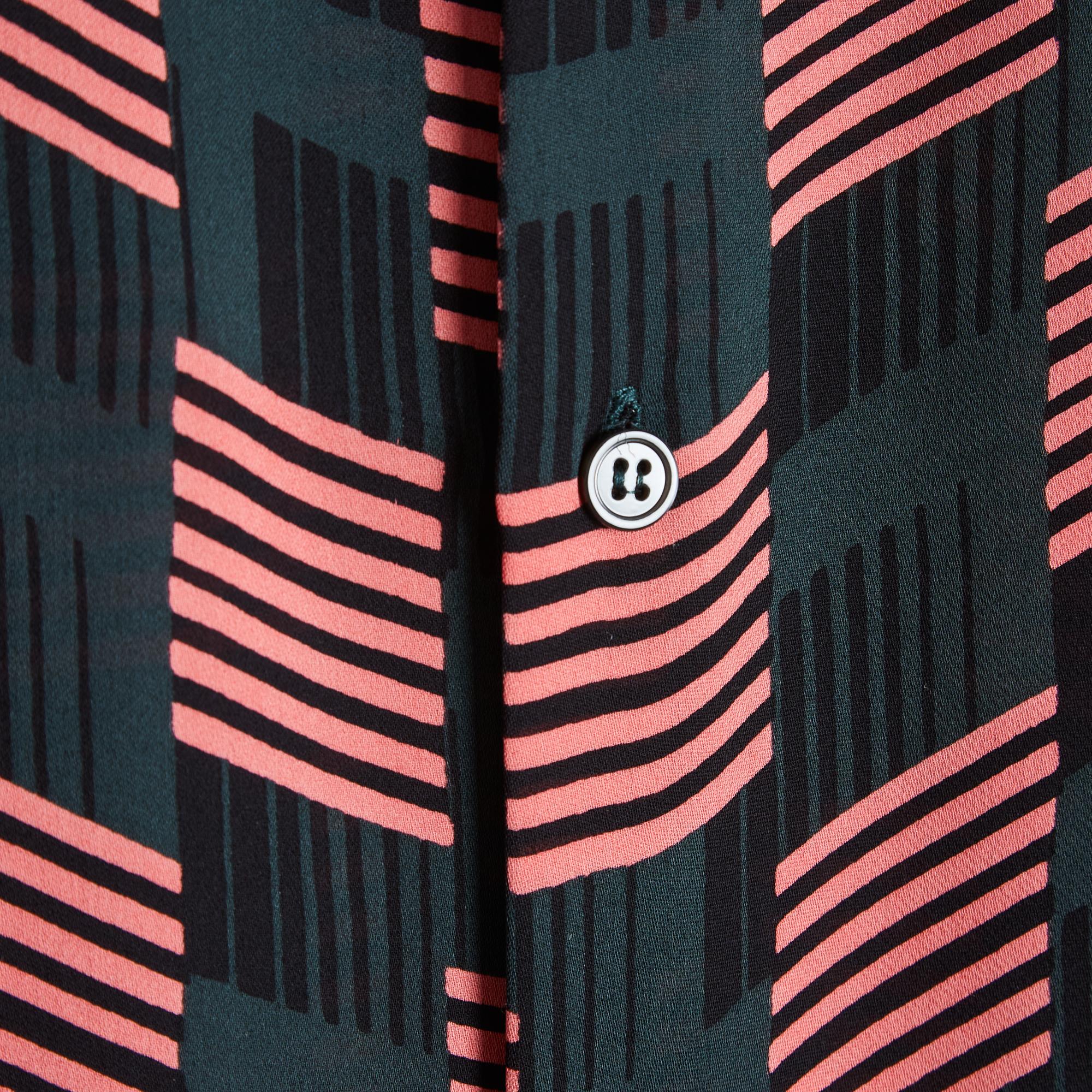 Blusa sobredimensionada Marni en ligero crepé de seda con estampado de cuadros en verde oscuro, rosa y negro, inspirada en los estilos africanos. Tiene cuello redondo, se abrocha en la espalda con siete botones y dos lazos, y las mangas son largas y